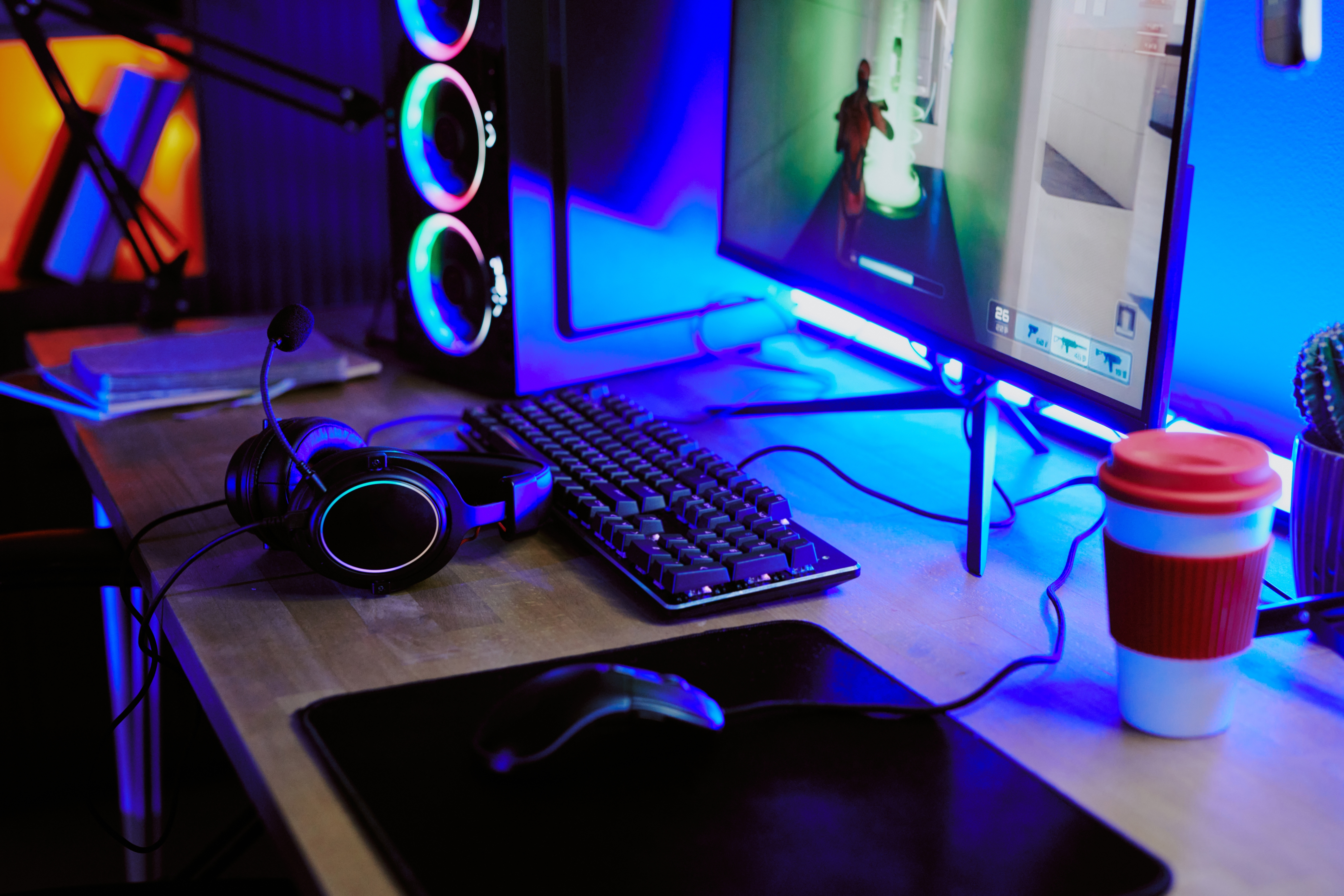 Spilloppsett med en skjerm som viser et skytterspill, tastatur, mus, hodetelefoner og en takeaway kaffekopp på skrivebordet