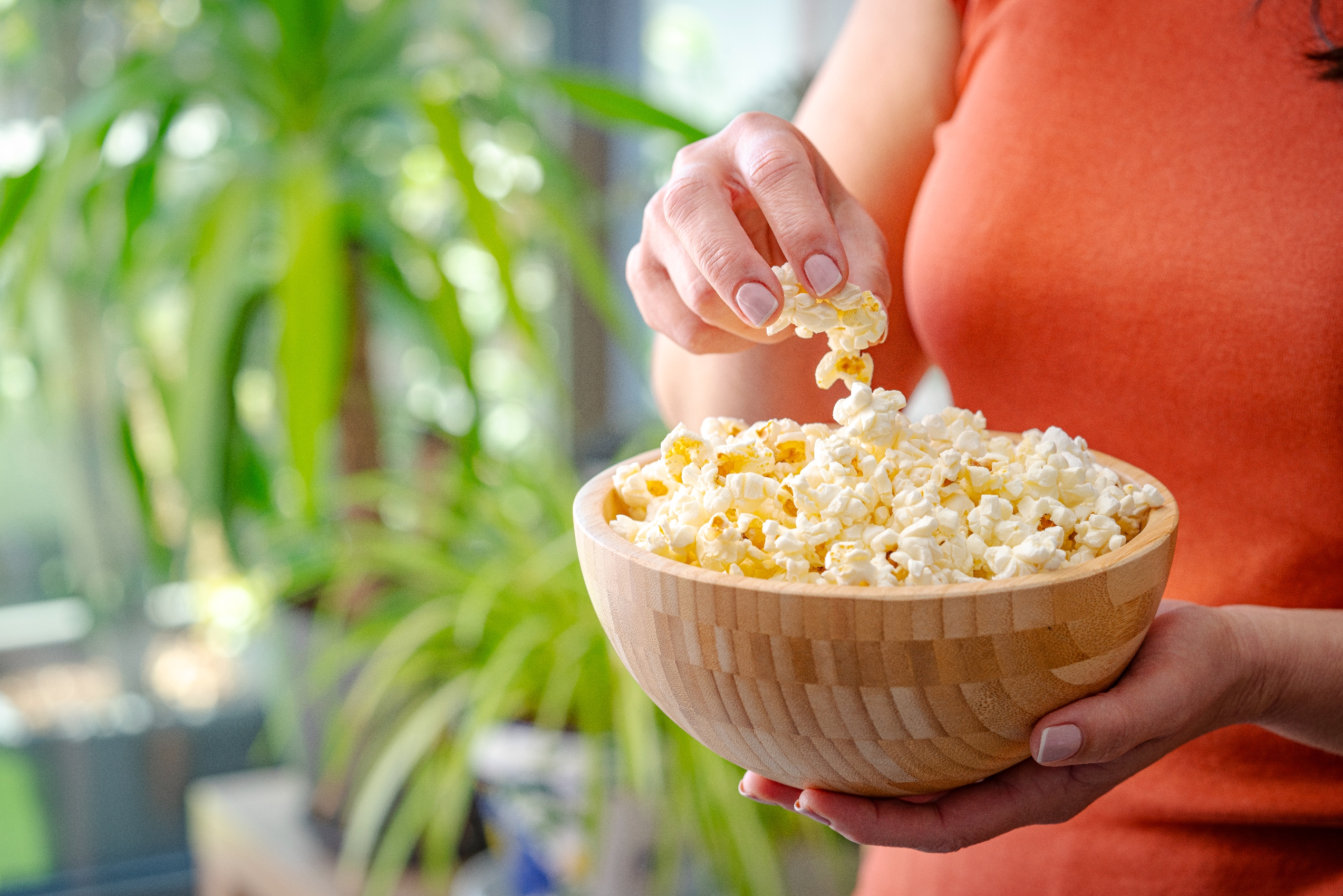 En person i en oransje skjorte holder en treskål med popcorn, og tar noen med hånden. En grønn plante er synlig i bakgrunnen