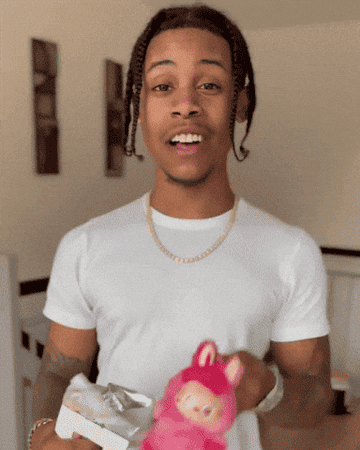 Person smiling, holding a gift box and a toy figure. They&#x27;re wearing a casual white t-shirt and a gold chain necklace indoors