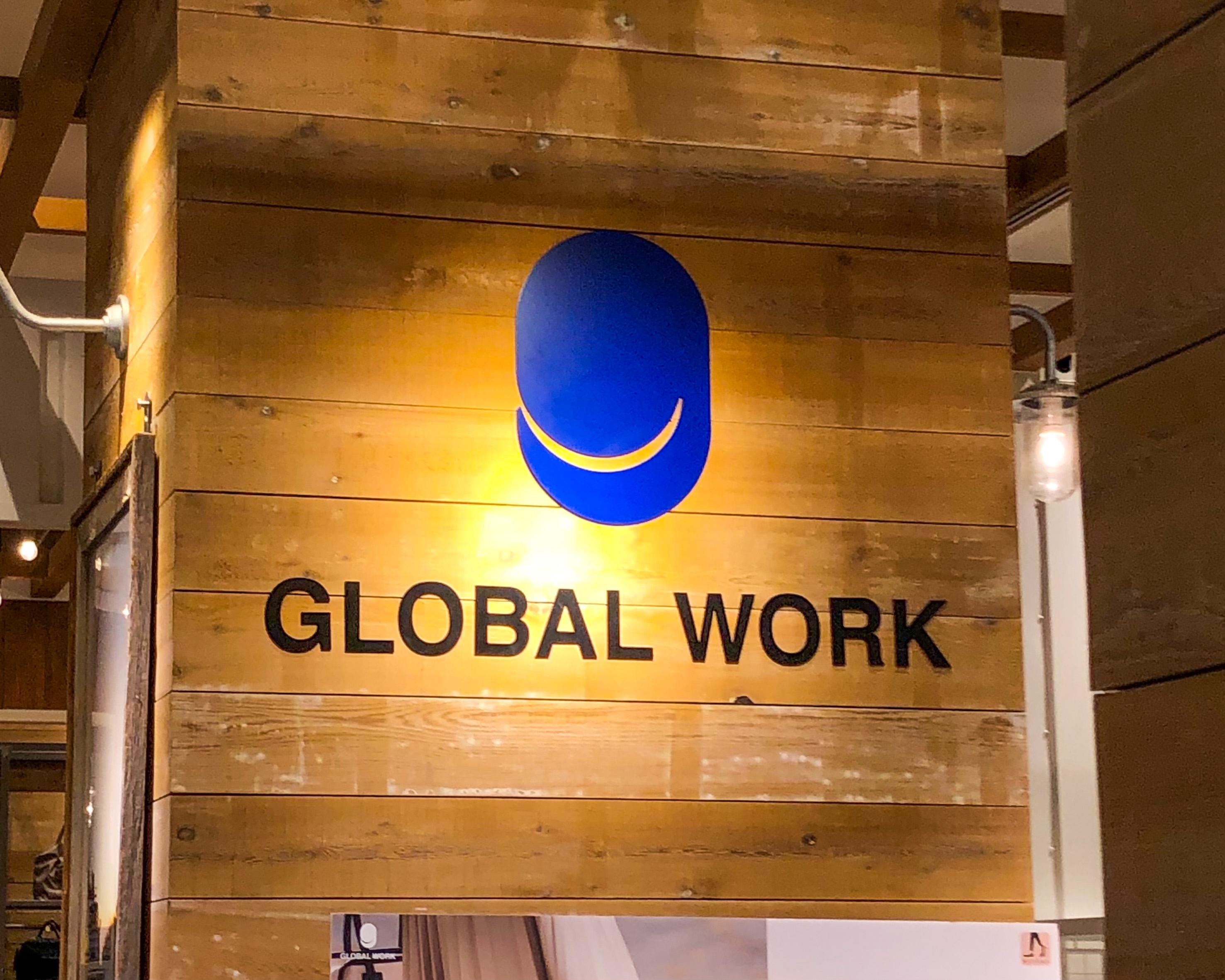 木製の壁に「GLOBAL WORK」というロゴが描かれている。ロゴデザインは青い楕円とその下の笑顔のライン。