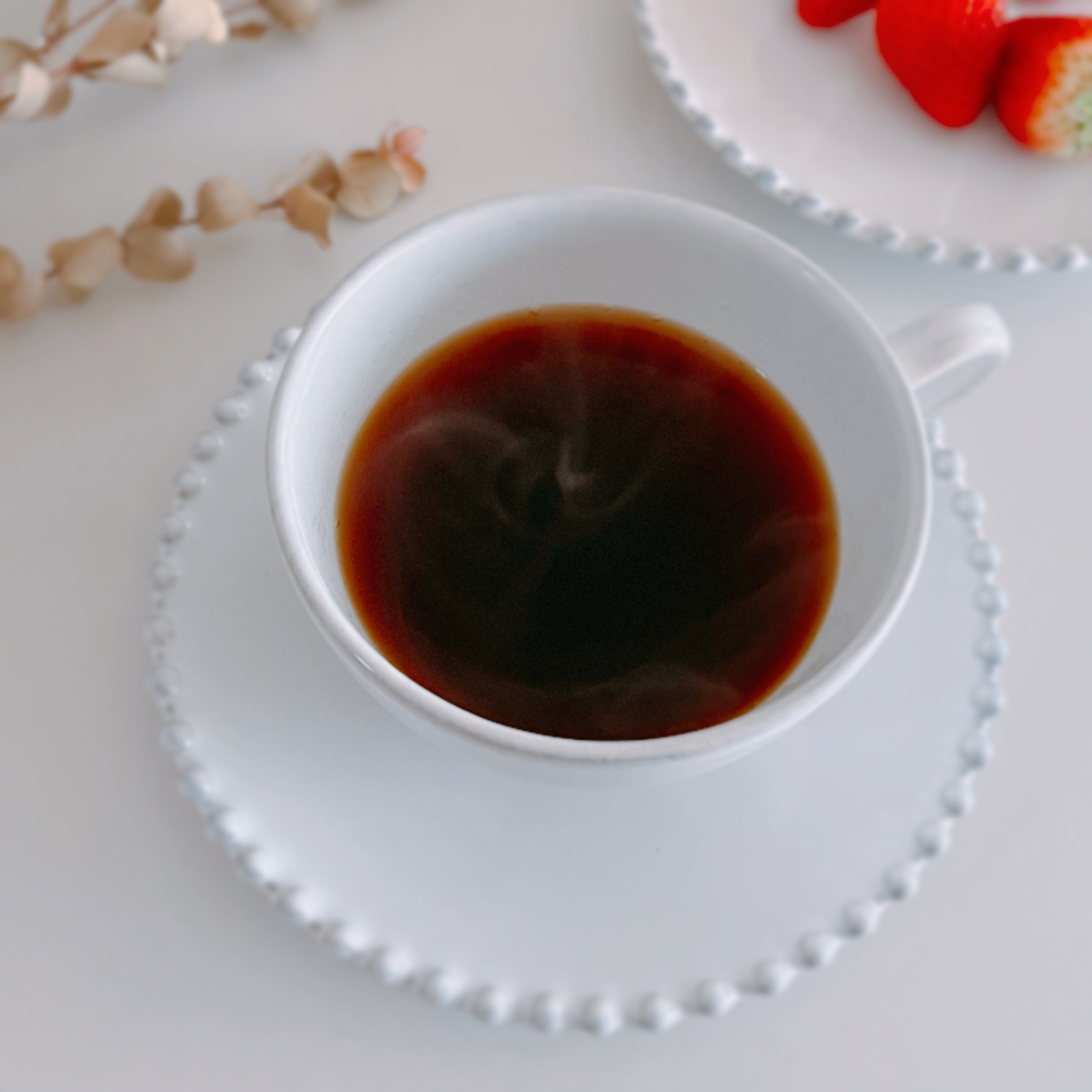 コーヒーカップに入った温かいコーヒーと、後ろにあるイチゴとケーキが白いプレートに載っています。