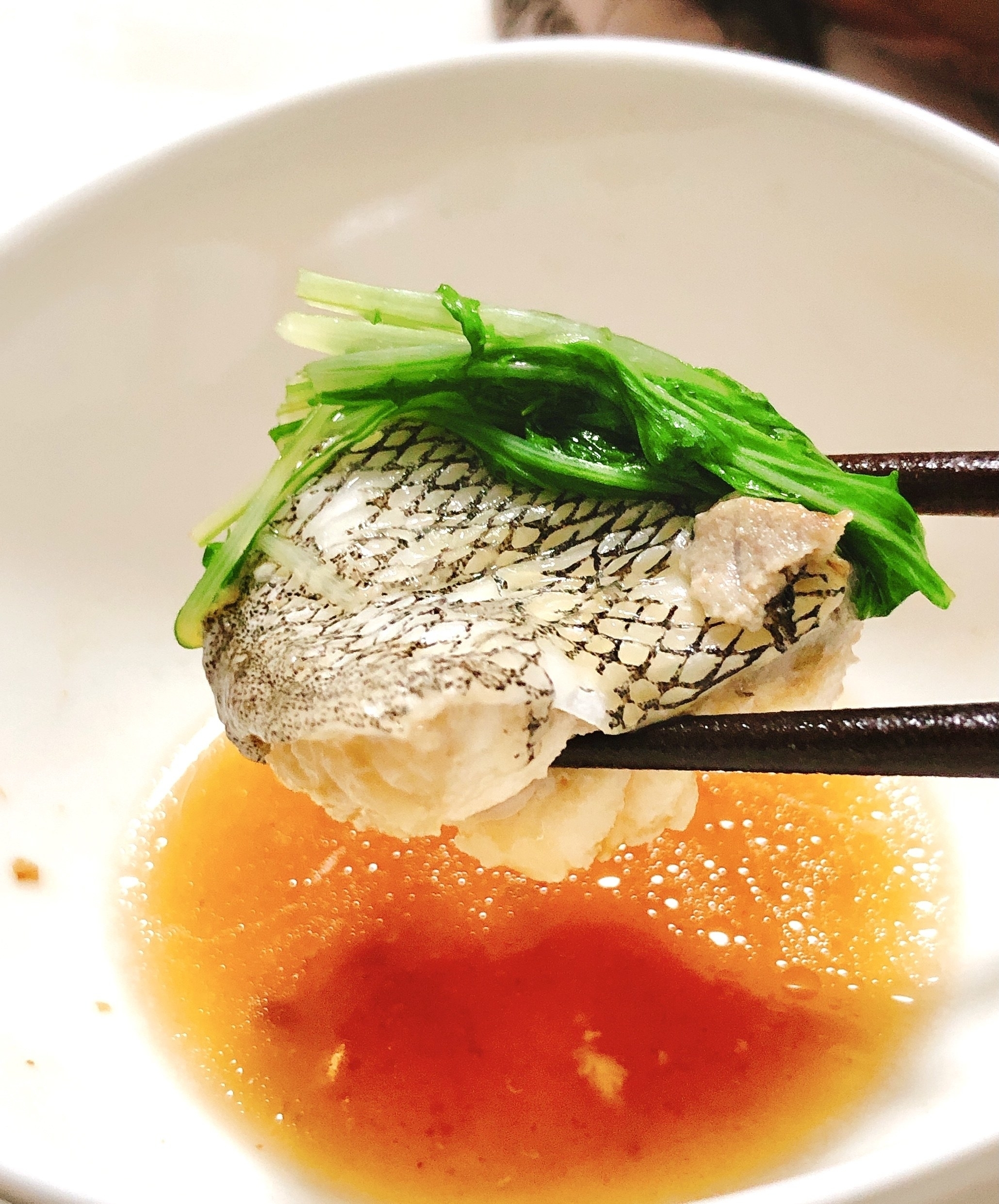 白いボウルに盛られたタレに、箸で持ち上げた魚の身と緑の野菜がつけられています。