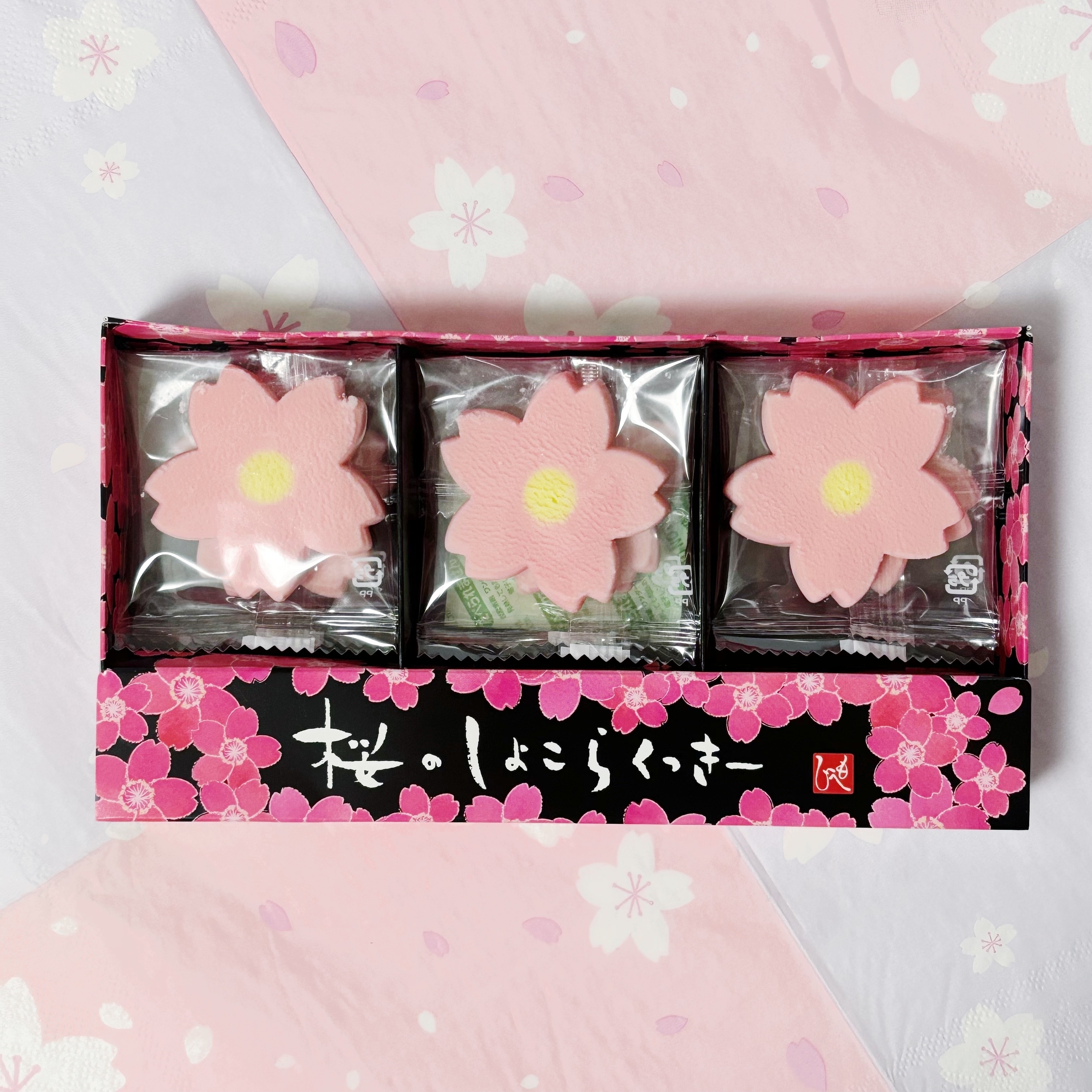 桜の形をした和菓子が3つ、包装された状態で箱に入っている。背景は花柄のデザイン。