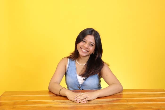Person smiling at a wooden table, wearing a denim vest and necklace, against a plain background