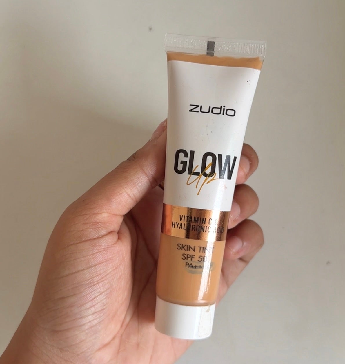 Hand holding a tube of Zudio Glow Up Vitamin C and Hyaluronic Skin Tint SPF 15, shown against a neutral background