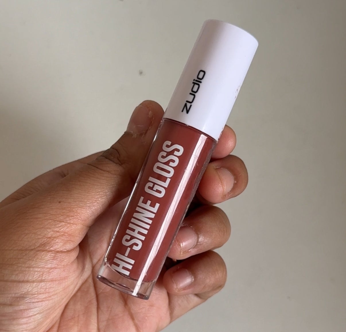 Person holds a tube of Hi-Shine Gloss with a white cap, labeled "Zudio."