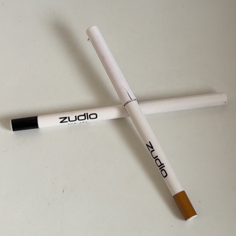 Two white eyeliner pencils cross each other on a light surface. One pencil has a black cap, and the other has a brown cap