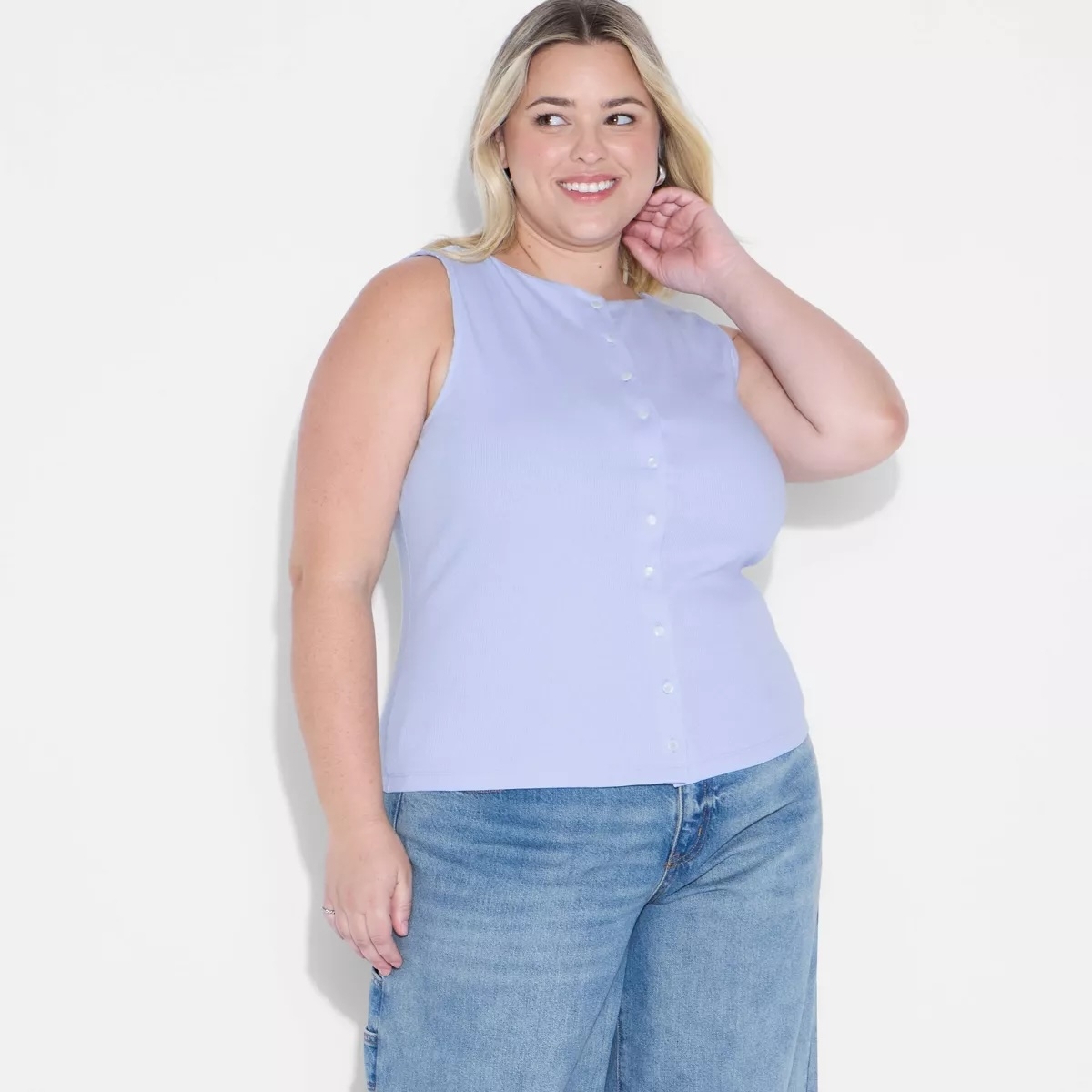 Person wearing a sleeveless button-up top and jeans, posing confidently