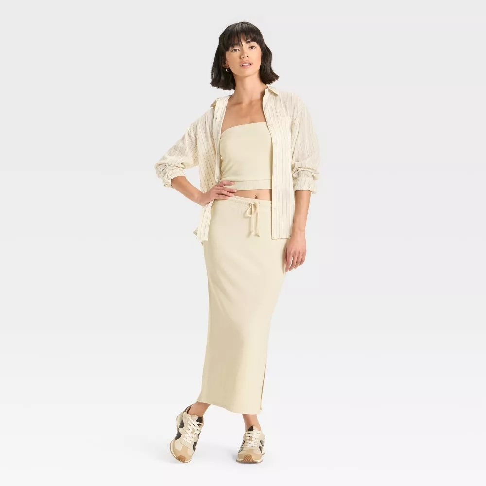Person in a casual beige outfit with a long skirt, tube top, and open button-down shirt, wearing sneakers