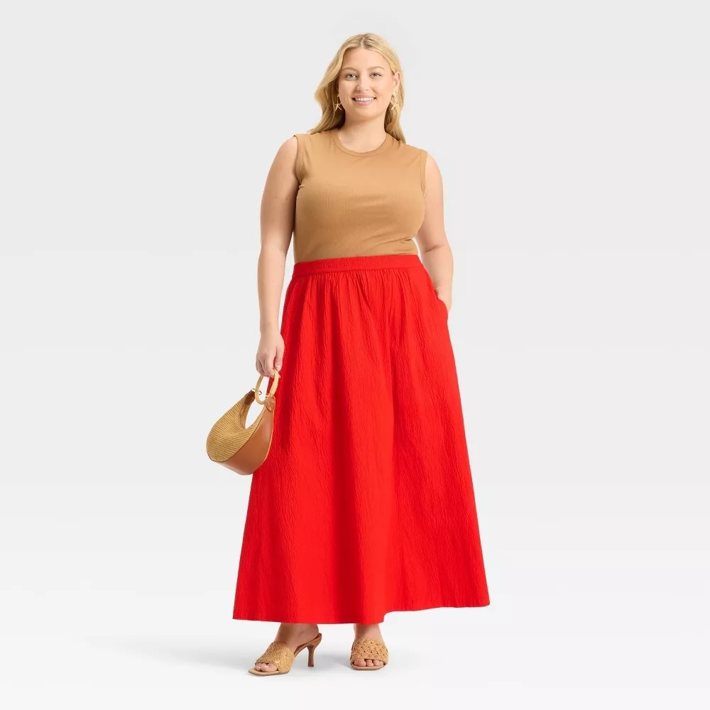 A person poses in a sleeveless top and long skirt, holding a handbag. Their outfit is stylish and casual, suitable for a shopping context