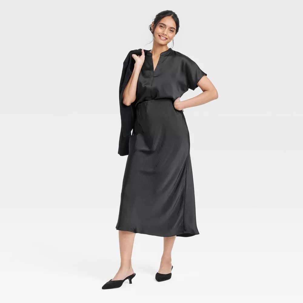 Person wearing a stylish, flowing black midi dress with short sleeves, holding a jacket over one shoulder, and black shoes