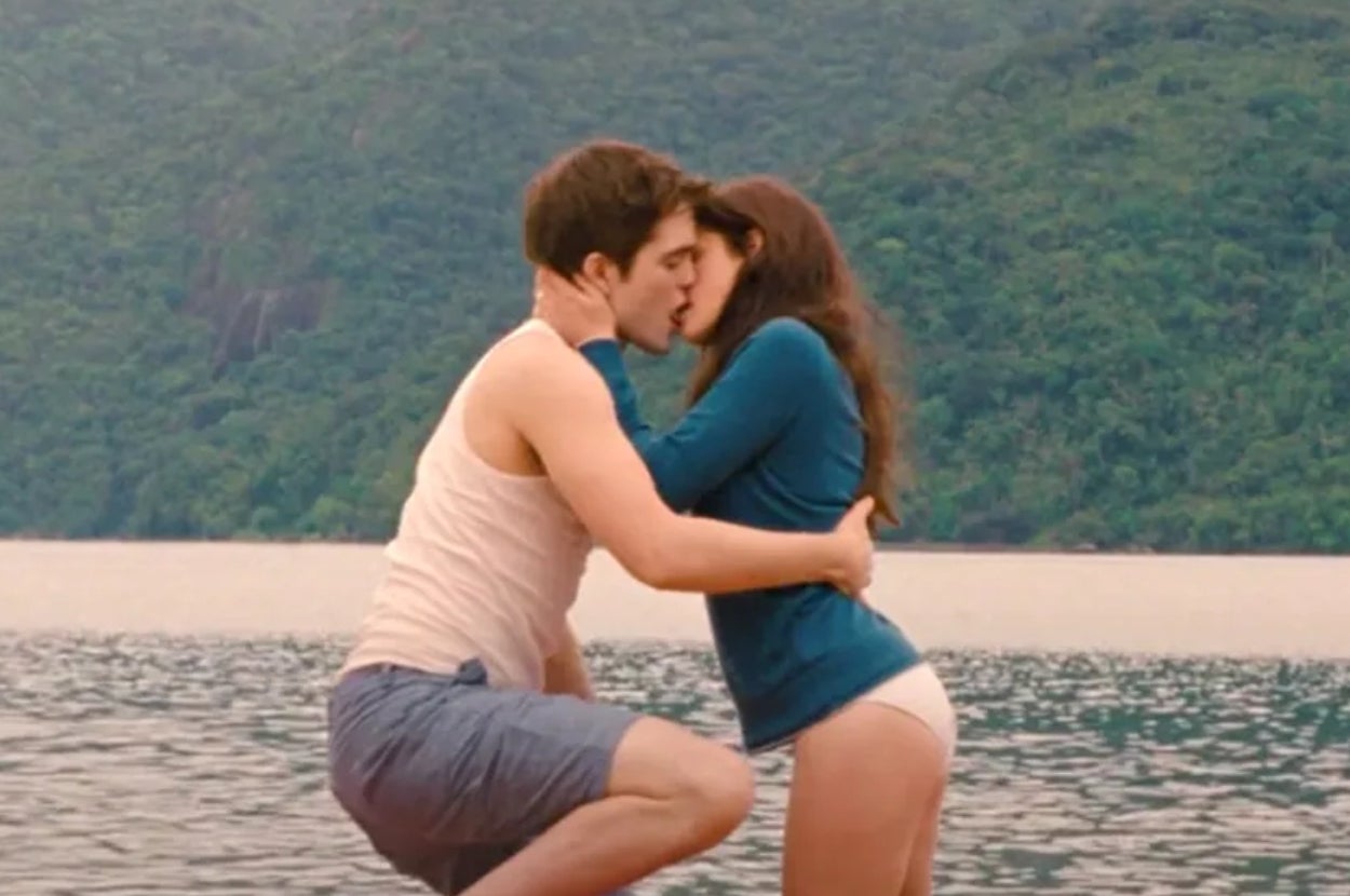 A couple kisses by a serene lakeside, surrounded by lush greenery. They are both dressed casually in light summer clothes
