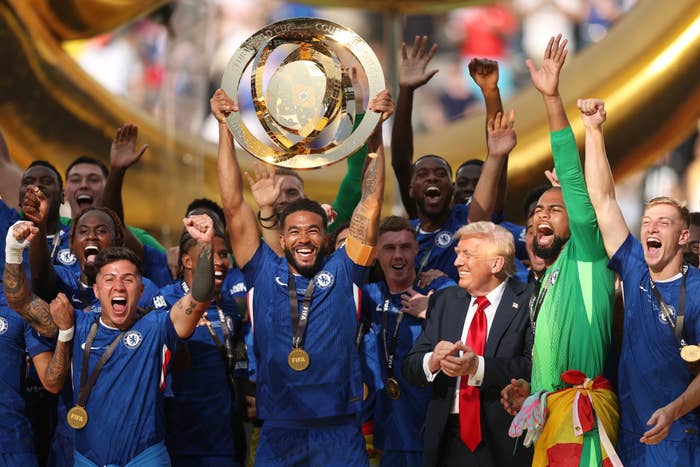Soccer team celebrates with a trophy; players are in blue jerseys. A man in a suit stands among them, cheering