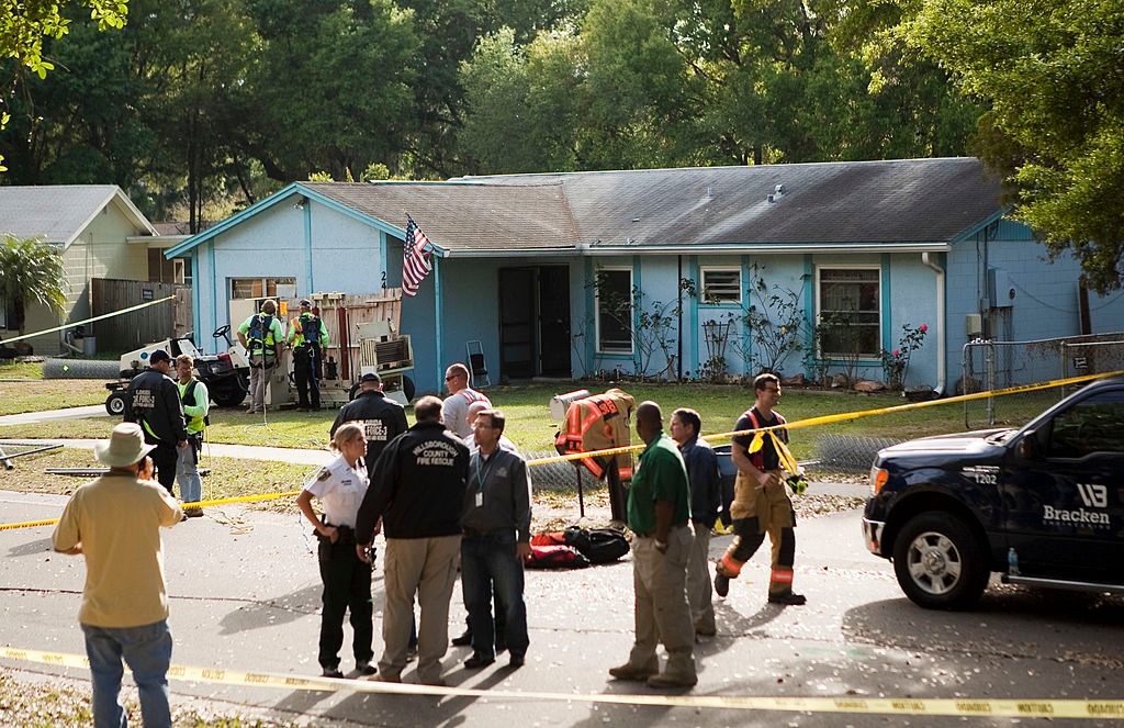 Emergency workers and officials gather outside a suburban home with an American flag, surrounded by caution tape and utility vehicles