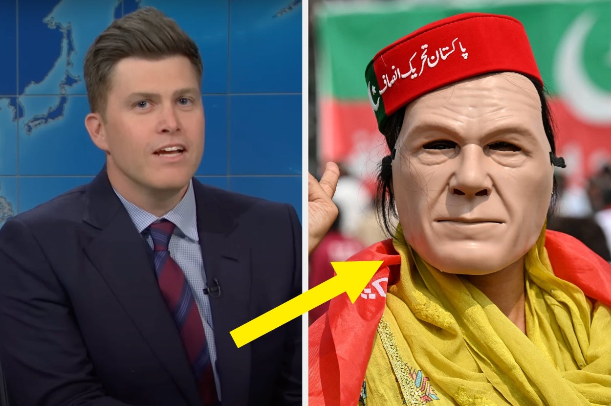 Person on a news set next to a protester holding a mask that seemingly resembles a public figure, with a yellow arrow pointing at the mask