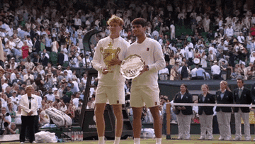 Two tennis players in athletic attire hold a trophy and plate on a tennis court, surrounded by a crowd celebrating their achievement