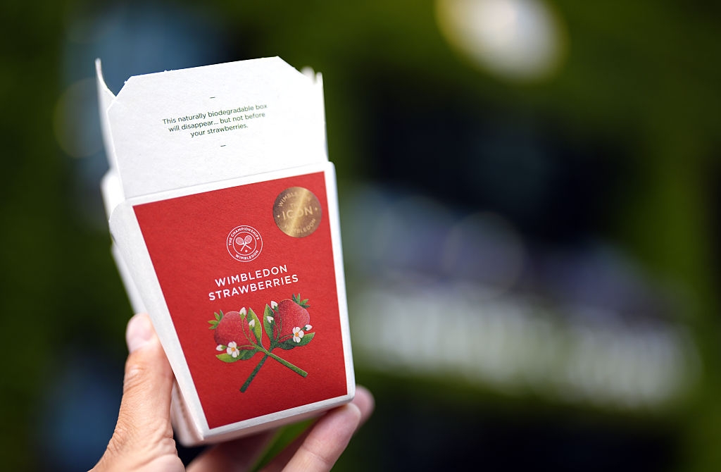 Hand holding a Wimbledon-branded box of strawberries with a special badge, against a blurred background