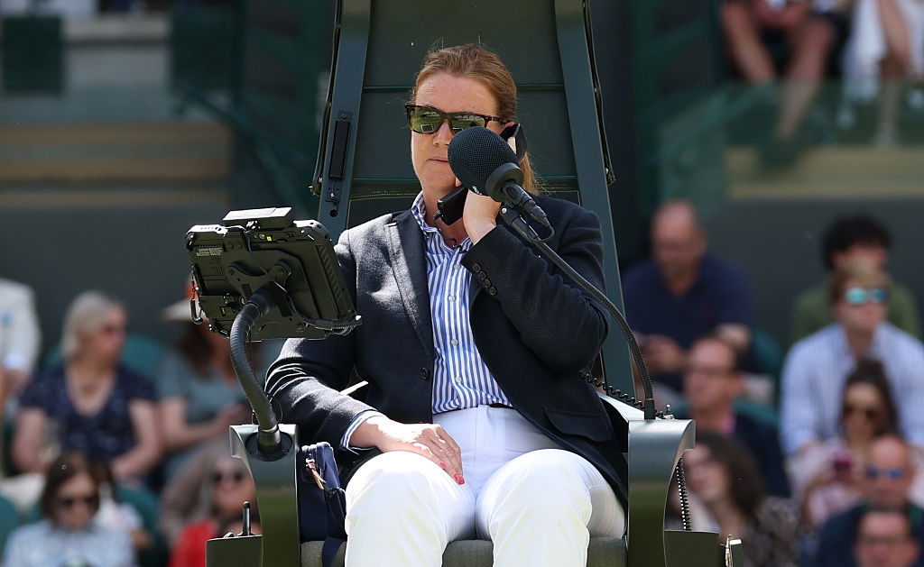 A tennis umpire in a blazer and sunglasses sits in a chair on a court, focused and holding a microphone, with spectators in the background