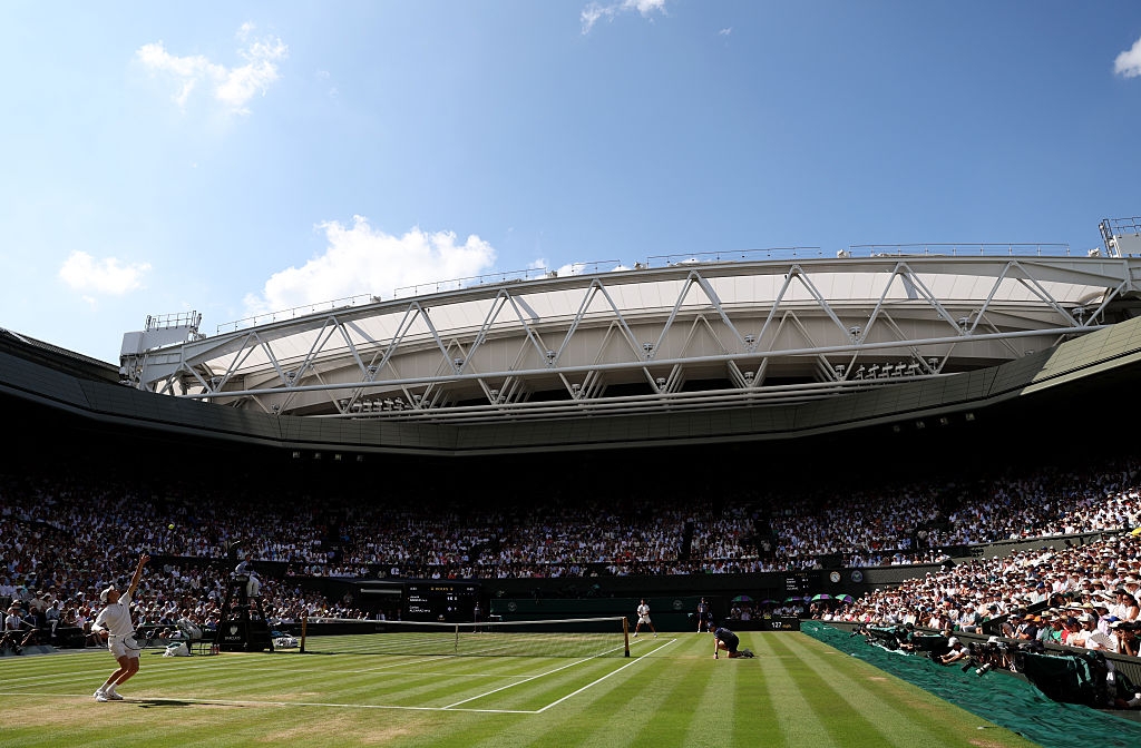 Tennis match in progress at Wimbledon with a large crowd. The stadium roof is open, revealing a clear sky. Players are positioned for a serve