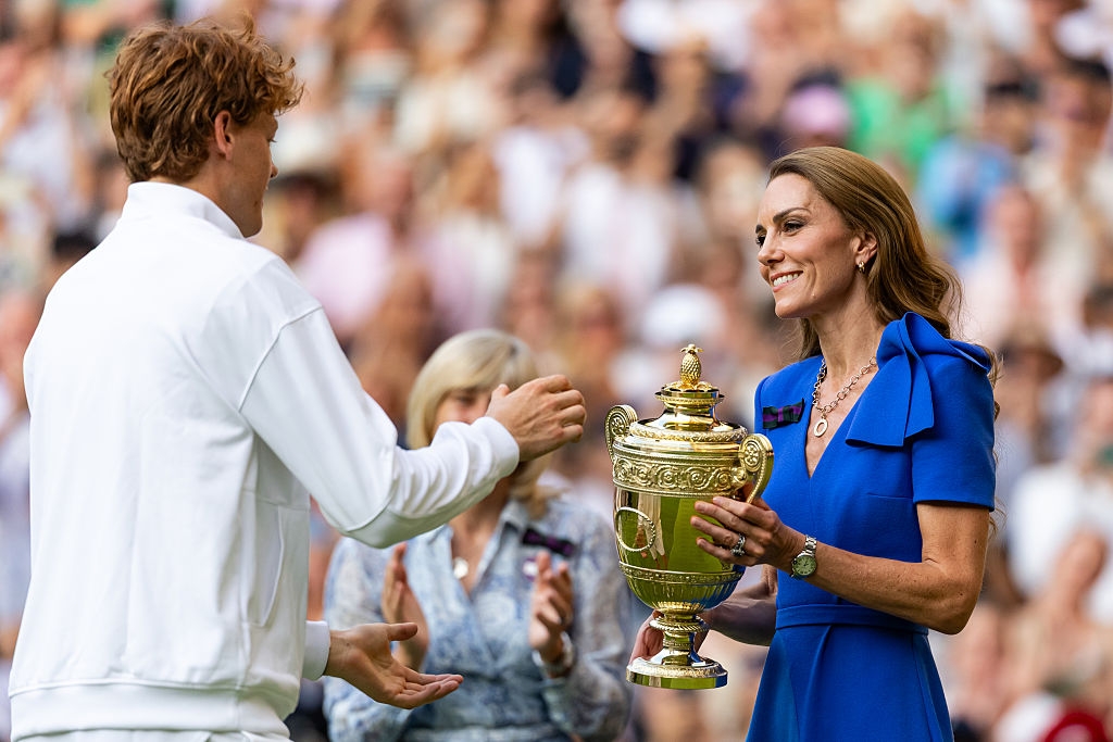 A woman in a blue dress presents a golden trophy to a tennis player on a court, with a crowd in the background