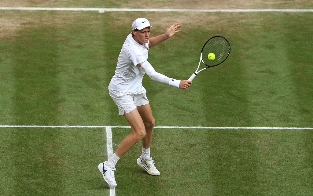 Tennis player on grass court, wearing a white outfit and cap, preparing to hit a backhand shot