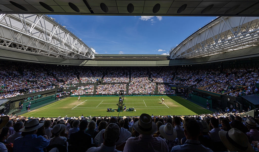 A crowded tennis stadium with players in action on the court. Spectators fill the seats under a retractable roof on a clear day