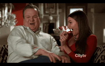 Two people on a couch burst into laughter, playfully surprised, one in a white sweater and the other in a red dress with long hair