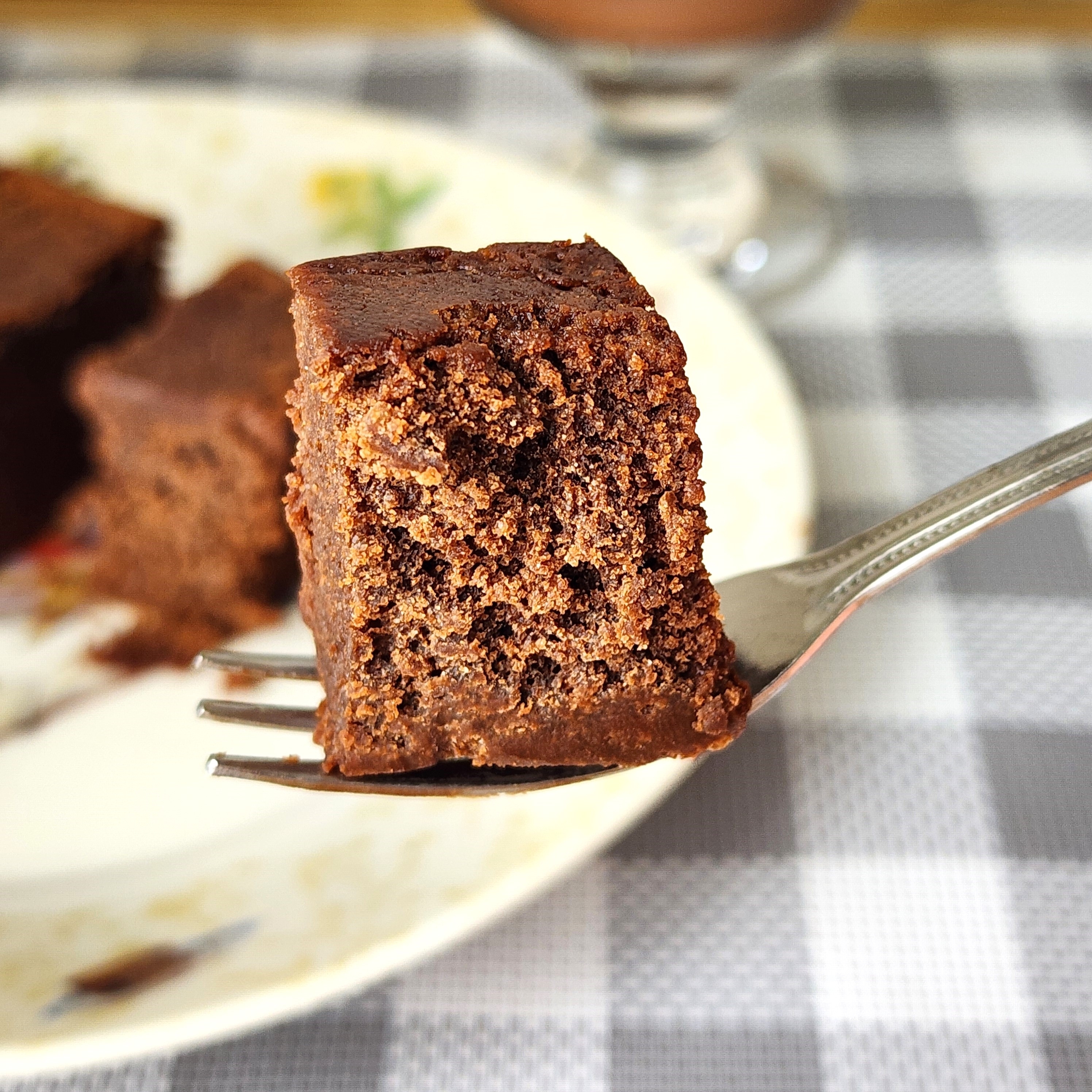 フォークにささった濃厚なチョコレートケーキの一切れ。背景にチェック柄のテーブルクロスと皿がある。