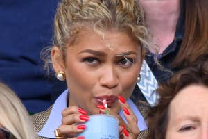 Person with styled hair sips from a cup with a straw at a public event, surrounded by others. Wears a blazer and bold nail color