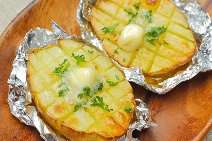 ホイルに包まれたハッセルバックポテトが木製皿に2つ。上にバターとハーブがトッピングされています。