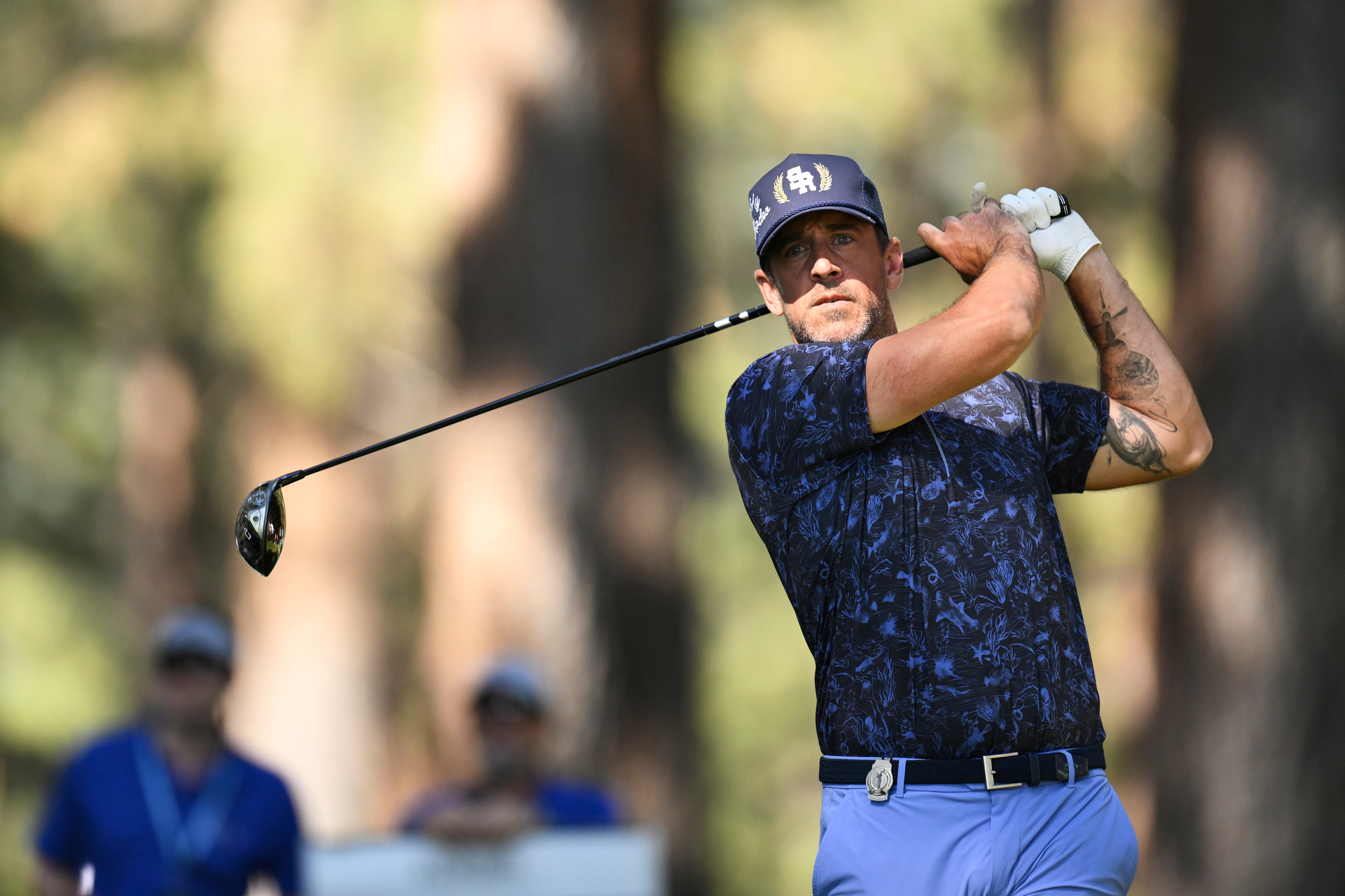 Man in golf attire, swinging a club on a golf course, focused on the shot. Background shows trees and spectators