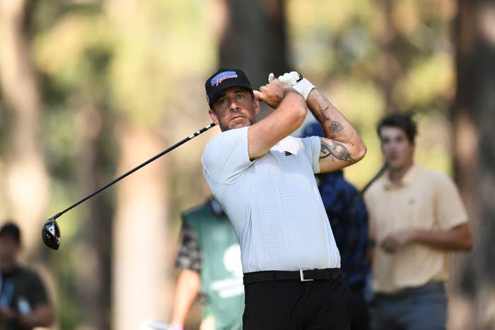 Celebrity golfer mid-swing on a golf course, wearing a polo shirt and cap, with spectators and trees in the background