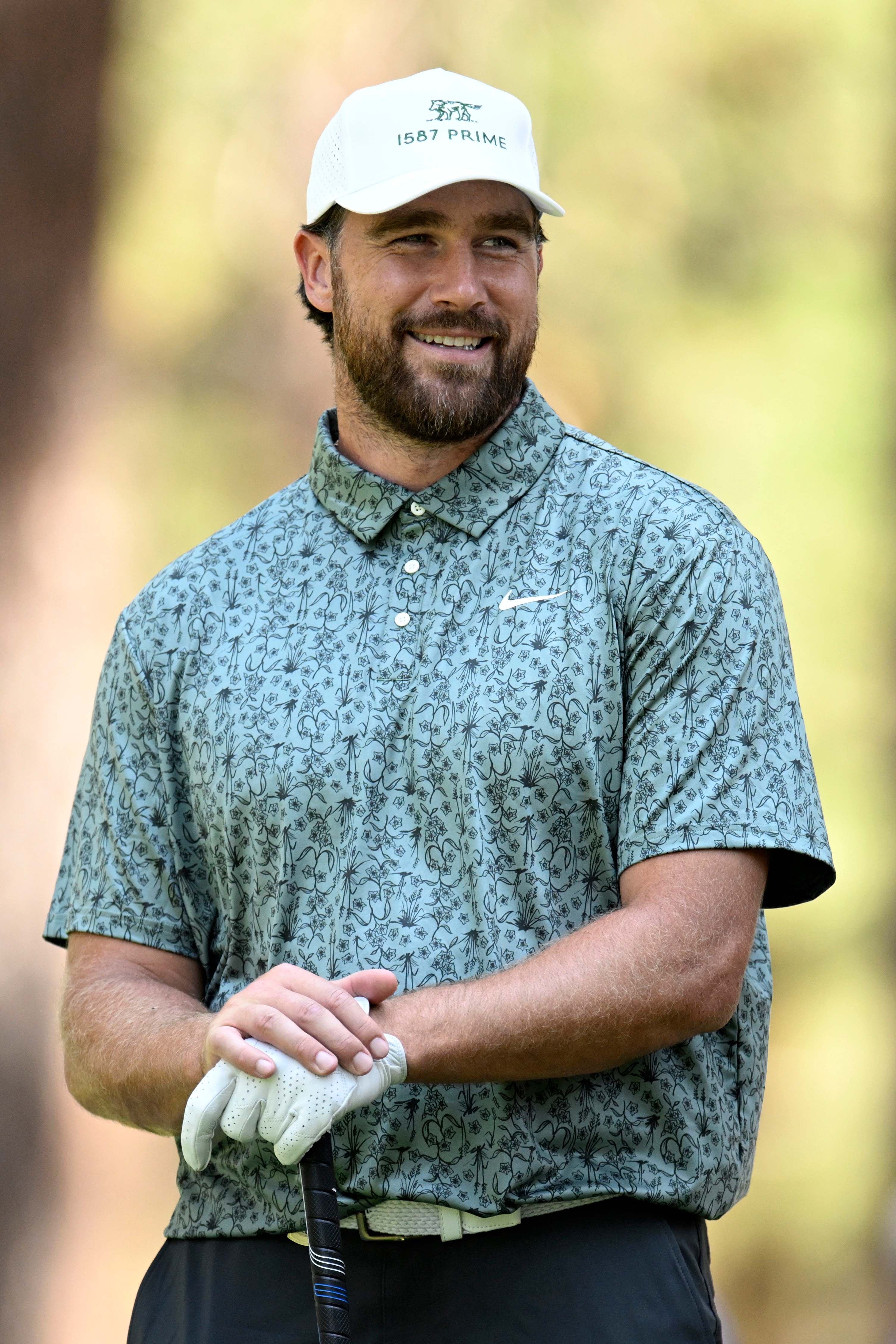 Person smiling, wearing a patterned shirt, cap, and holding a golf club, appears to be at a golf course