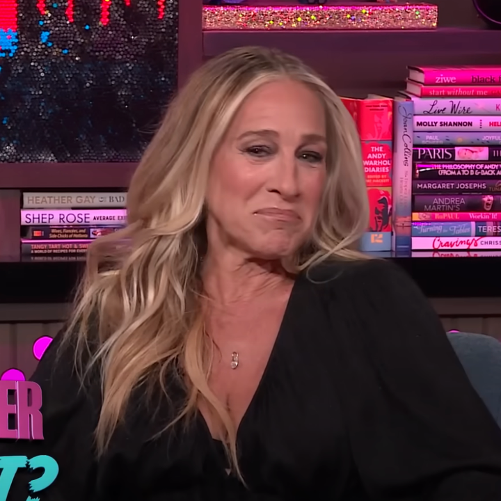 Person with long hair in a black outfit on a talk show set, surrounded by books and decorations