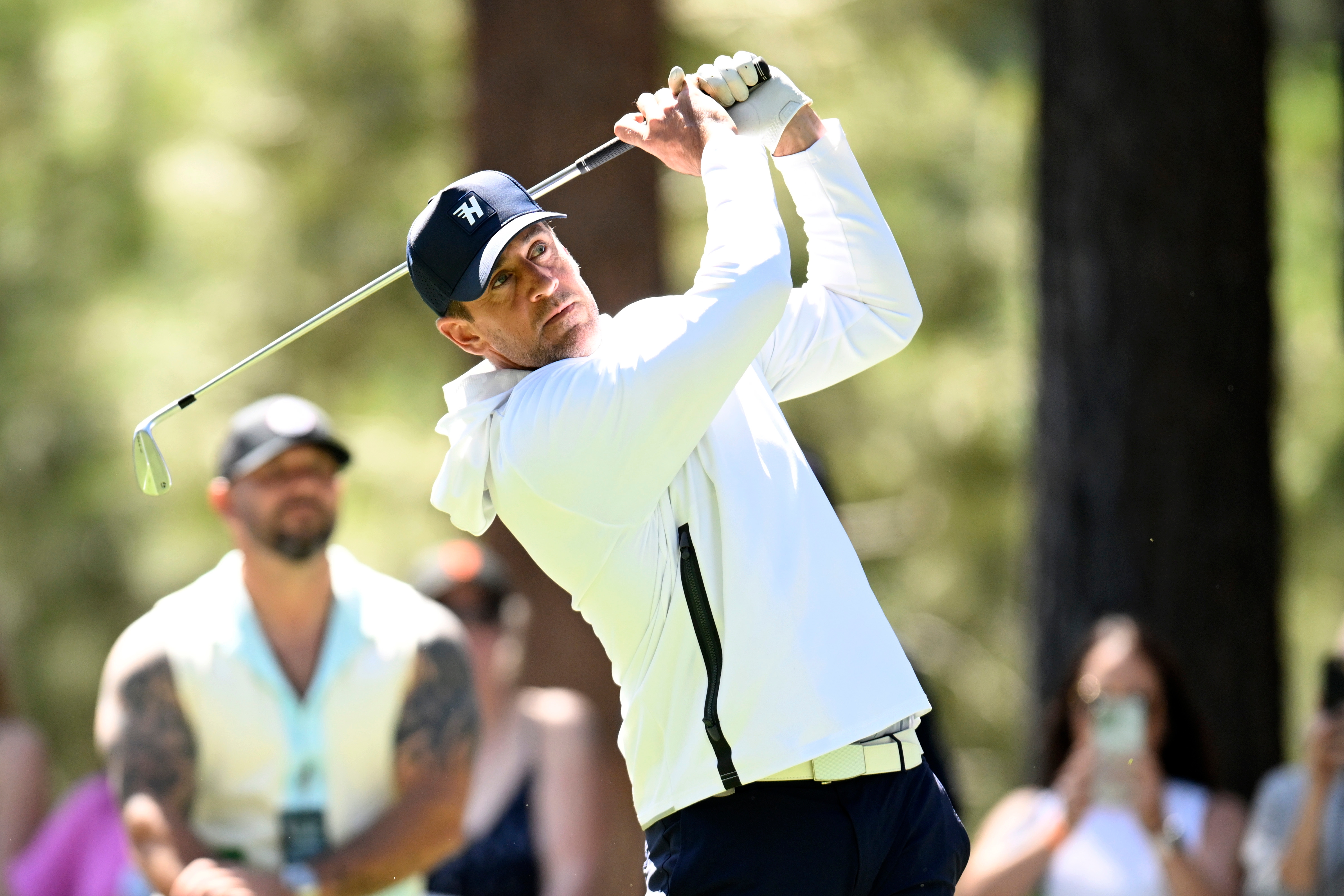 A golfer in a white top and cap swings a club on a course, surrounded by onlookers