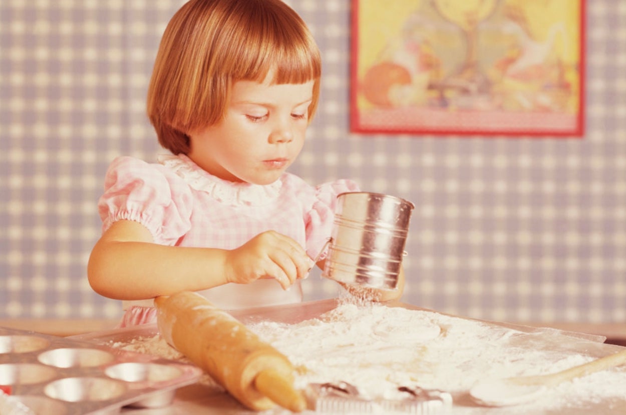 A vintage child in a pink dress bakes, using a sifter to sprinkle flour over a baking sheet, with a rolling pin and muffin tray nearby