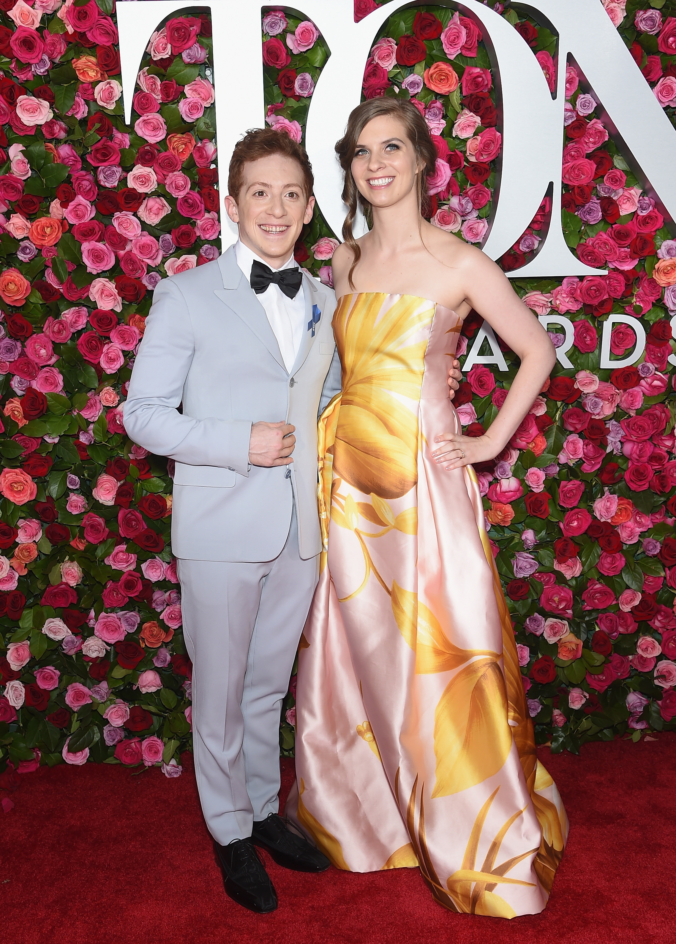 Two individuals on a red carpet; one in a light suit with a bow tie, the other in a floral strapless gown. Pink and white floral backdrop