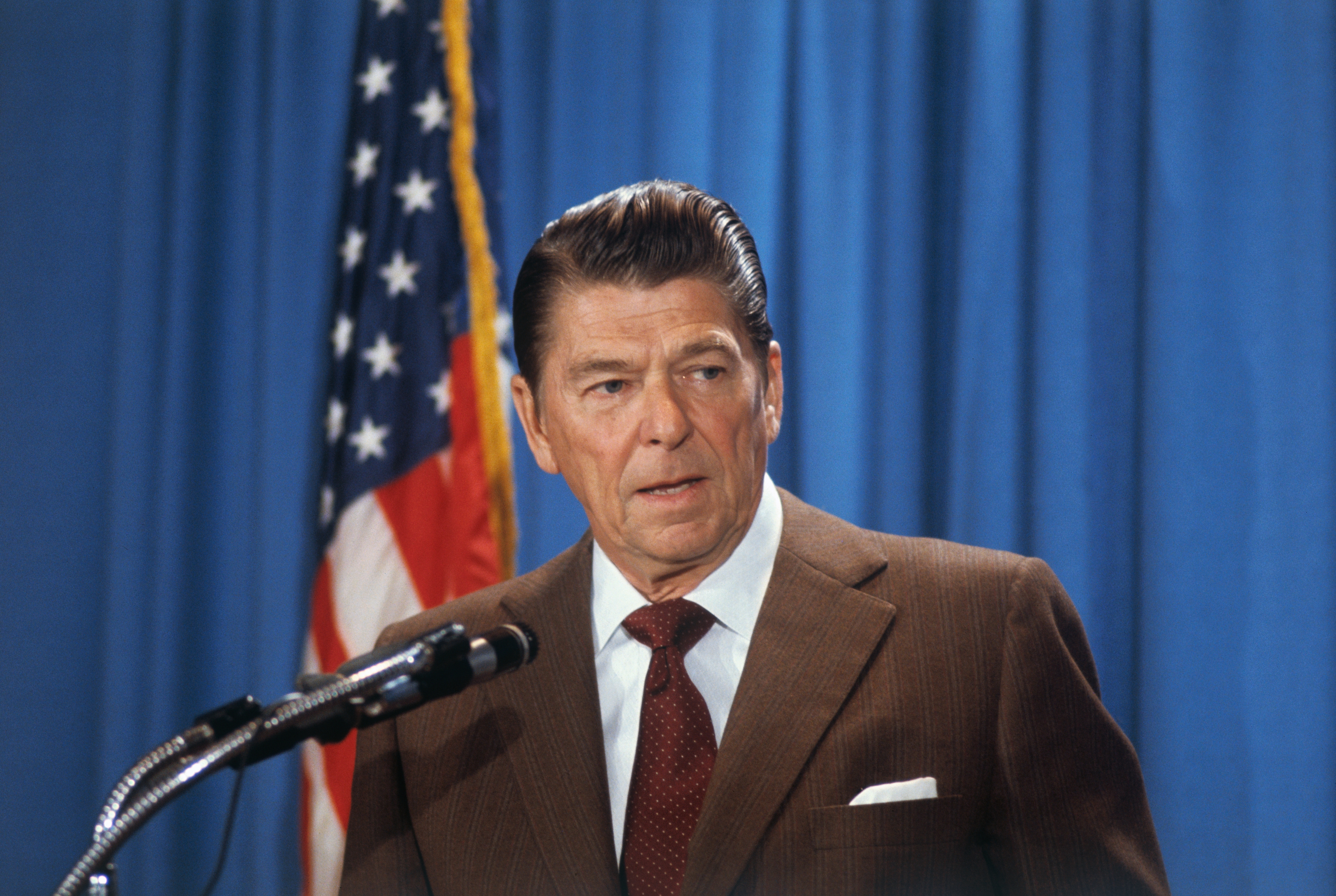 A person at a podium gives a speech in front of a U.S. flag backdrop. They wear a suit and tie