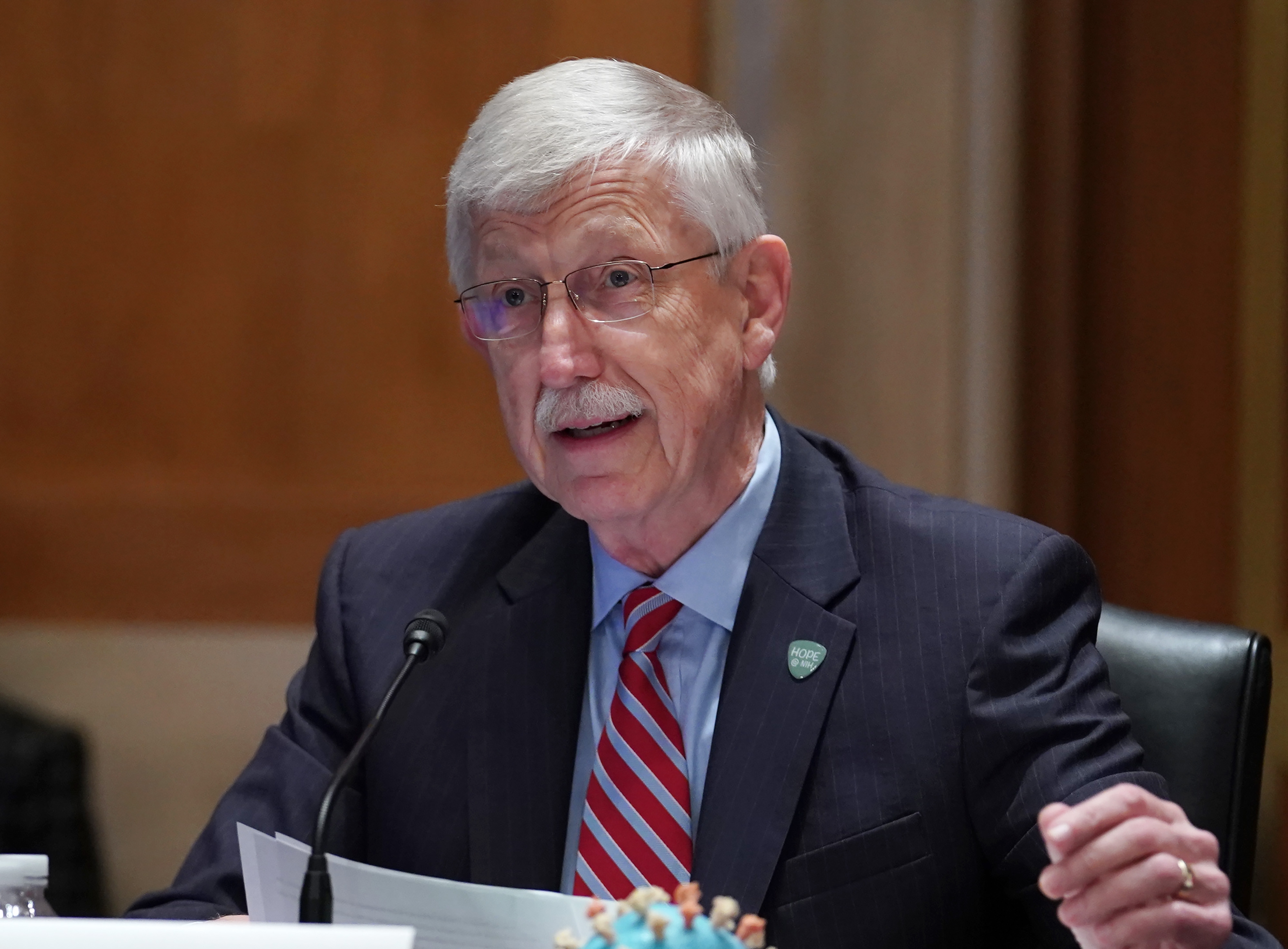 An older man in a suit and striped tie speaks at a conference table, holding papers, likely participating in a formal discussion or testimony