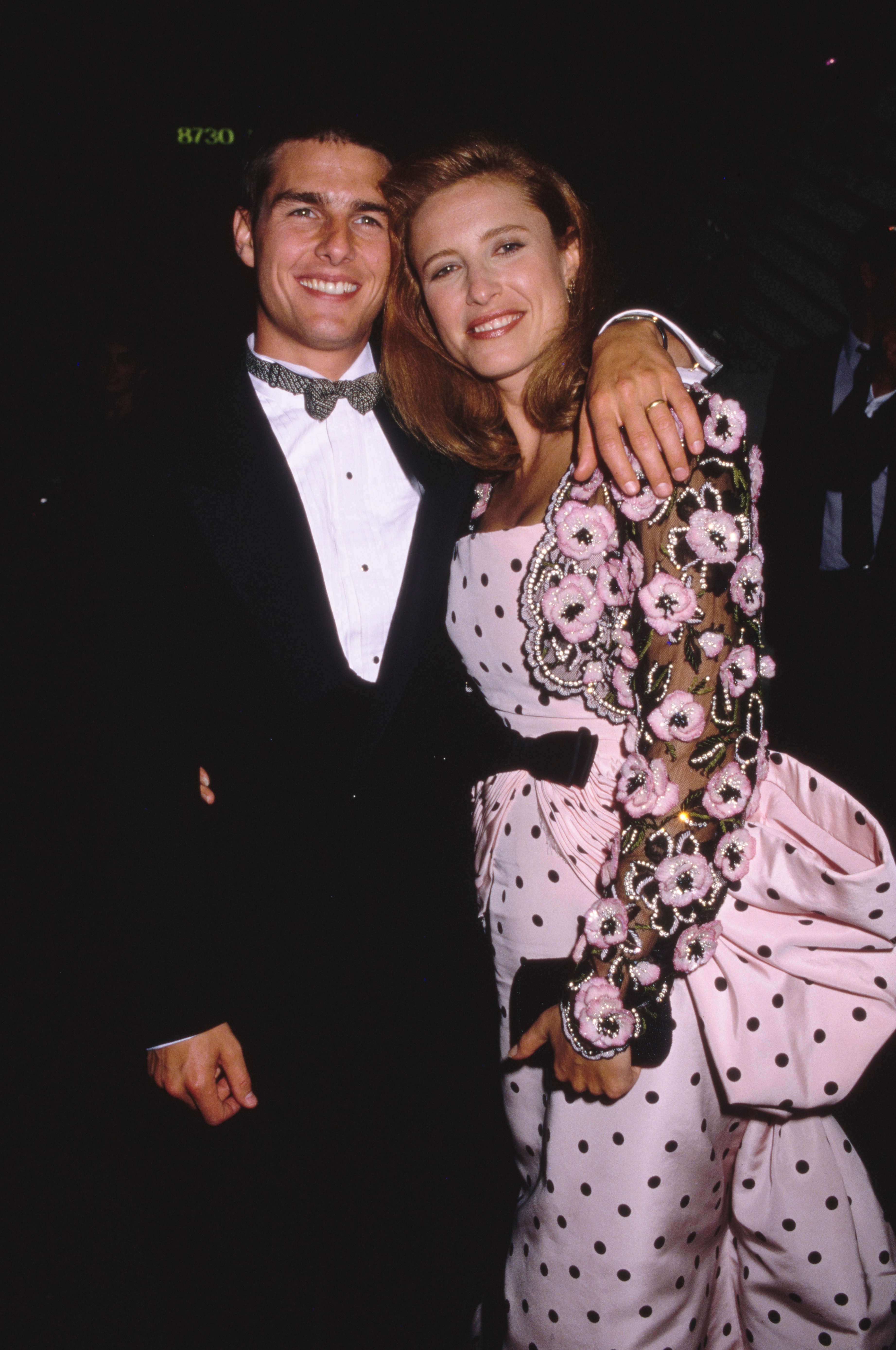 A man in a tuxedo with a bow tie and a woman in a floral and polka dot gown smile and pose at a formal event