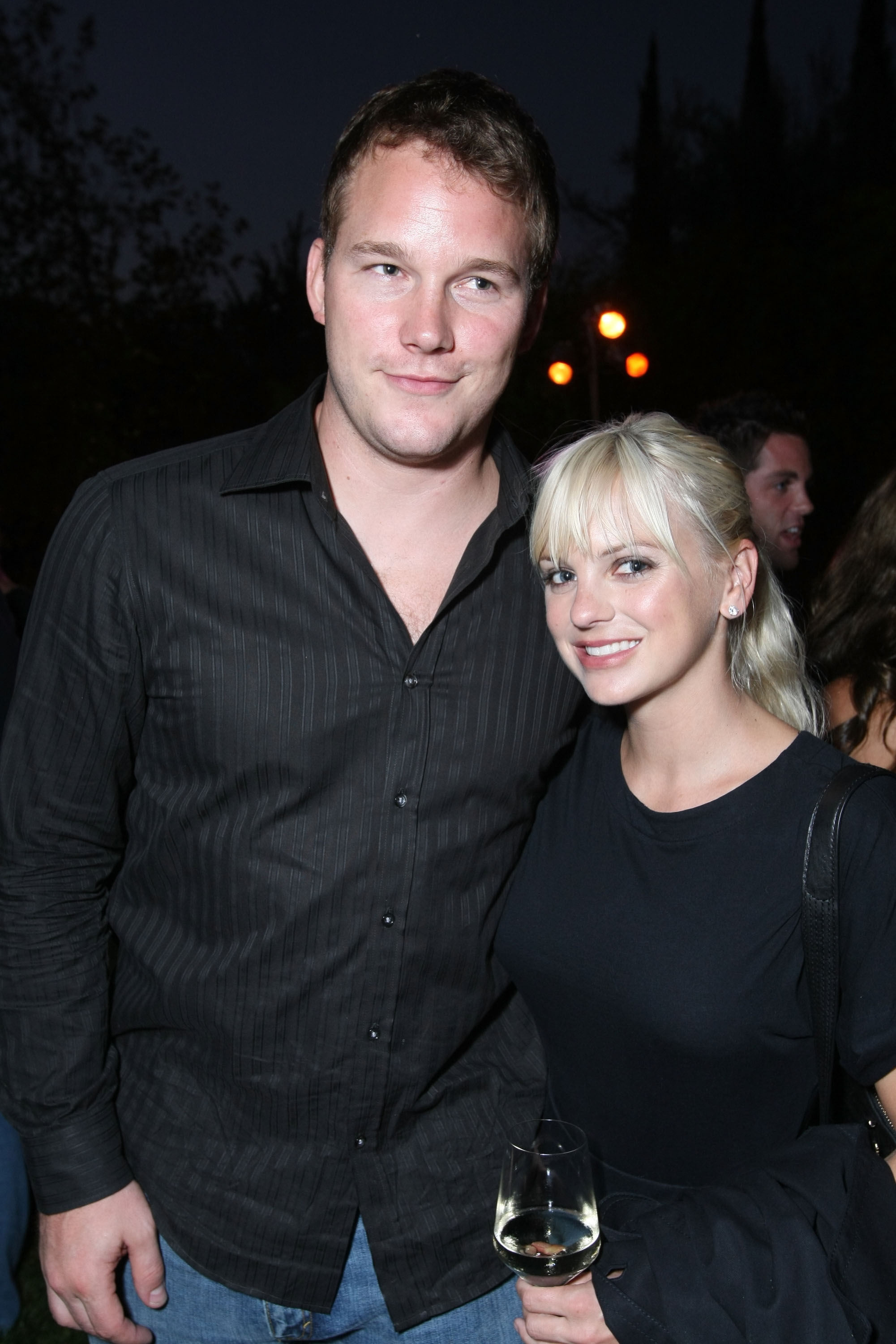 A man in a dark shirt and a woman in a black dress smile at a night event. She holds a glass of wine