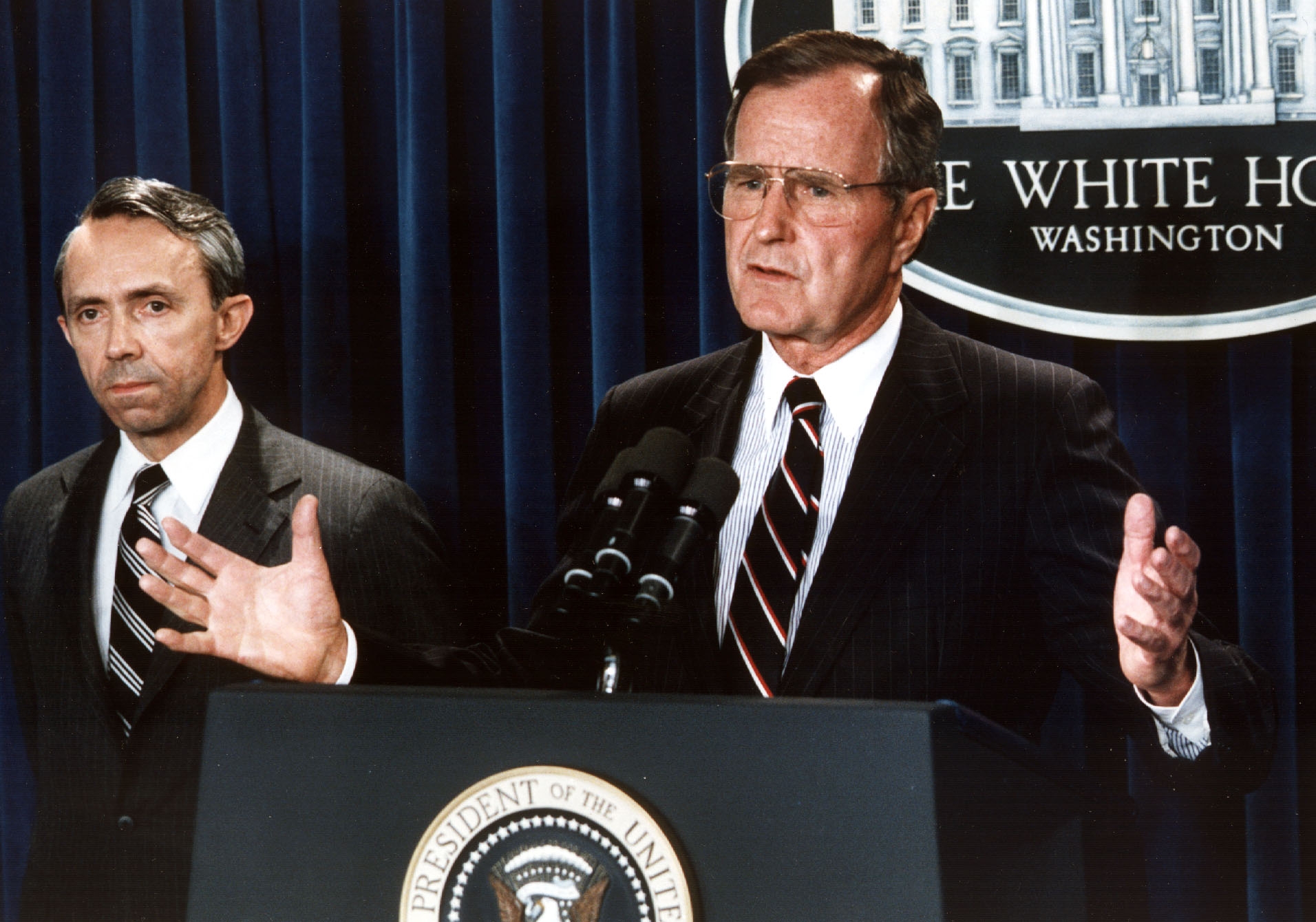 George H. Bush at a podium with a presidential seal, speaking to an audience, in a formal setting with a suit and tie