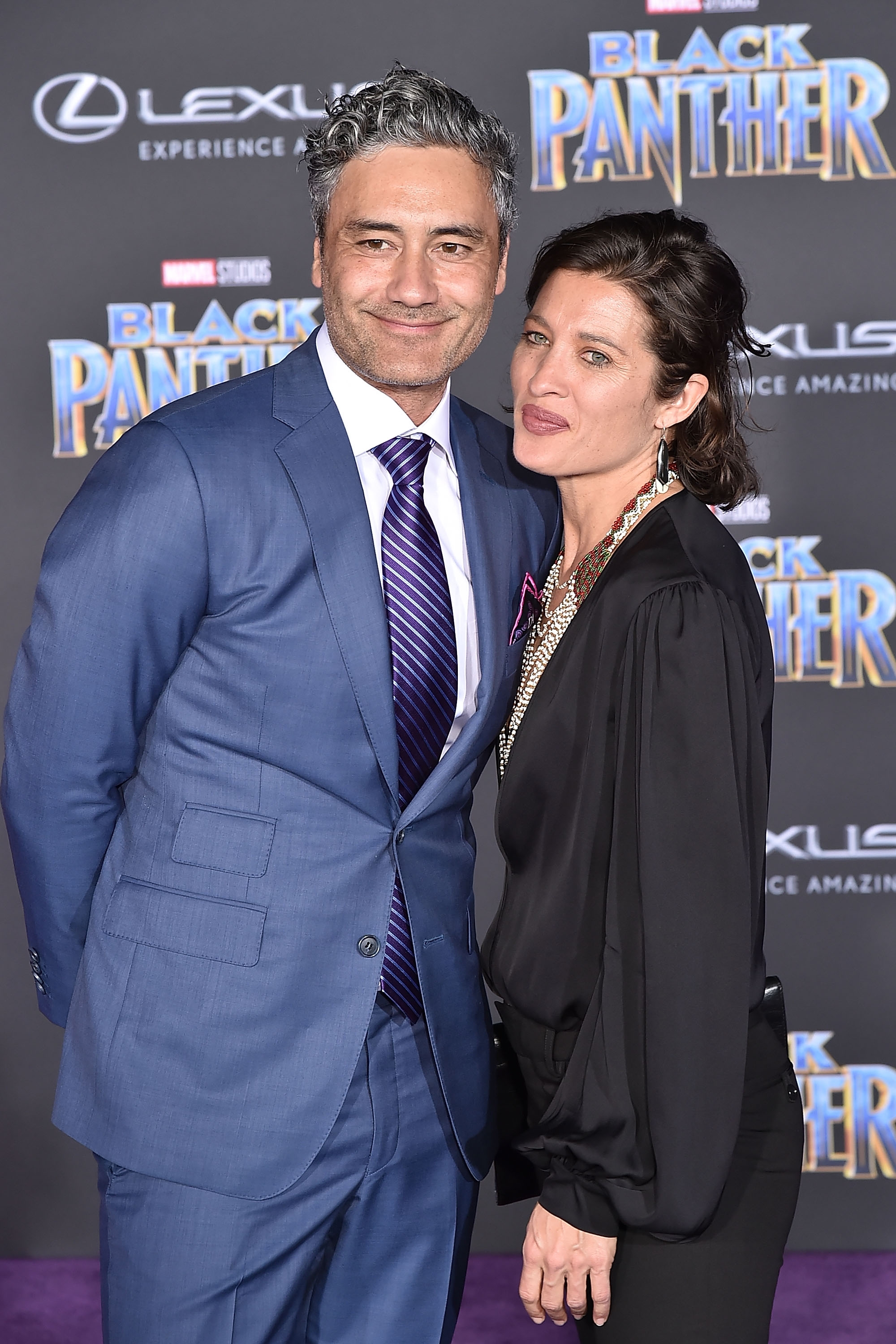 Two people on a red carpet. One is in a suit with a tie, and the other is in a long-sleeve blouse with necklace accessories, in front of a movie backdrop