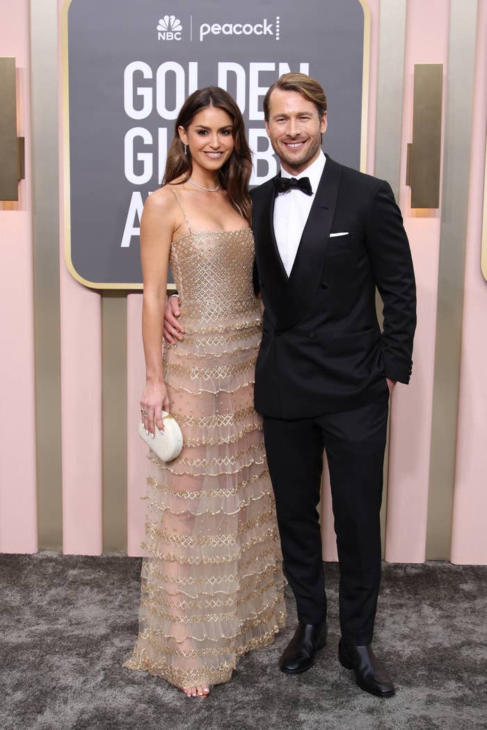 Two people on a red carpet; one wears a strapless, gold pattern dress with a clutch, the other a classic tuxedo. Golden Globe Awards backdrop visible