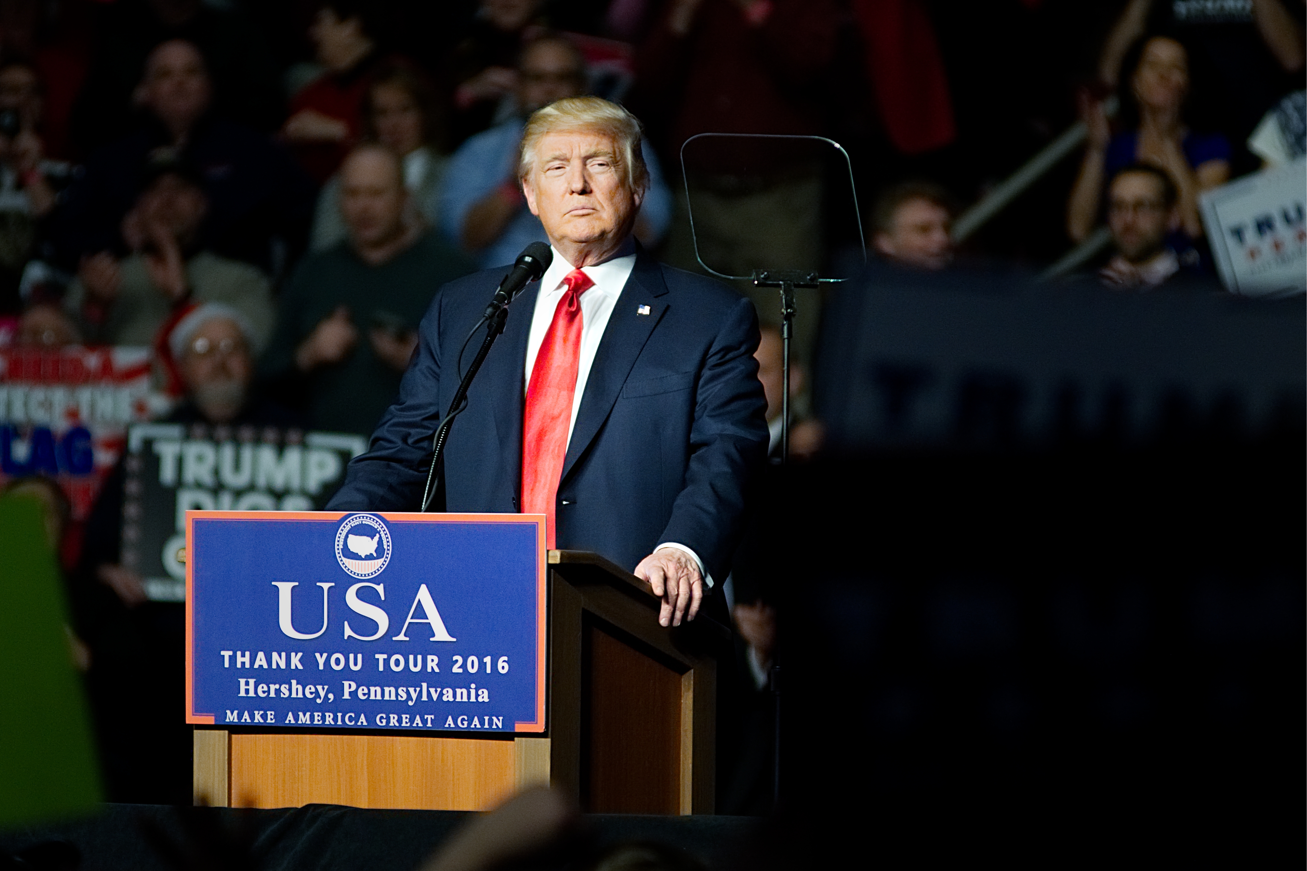 Person speaking at a podium with &quot;USA Thank You Tour 2016&quot; text. Background includes audience members and campaign signs