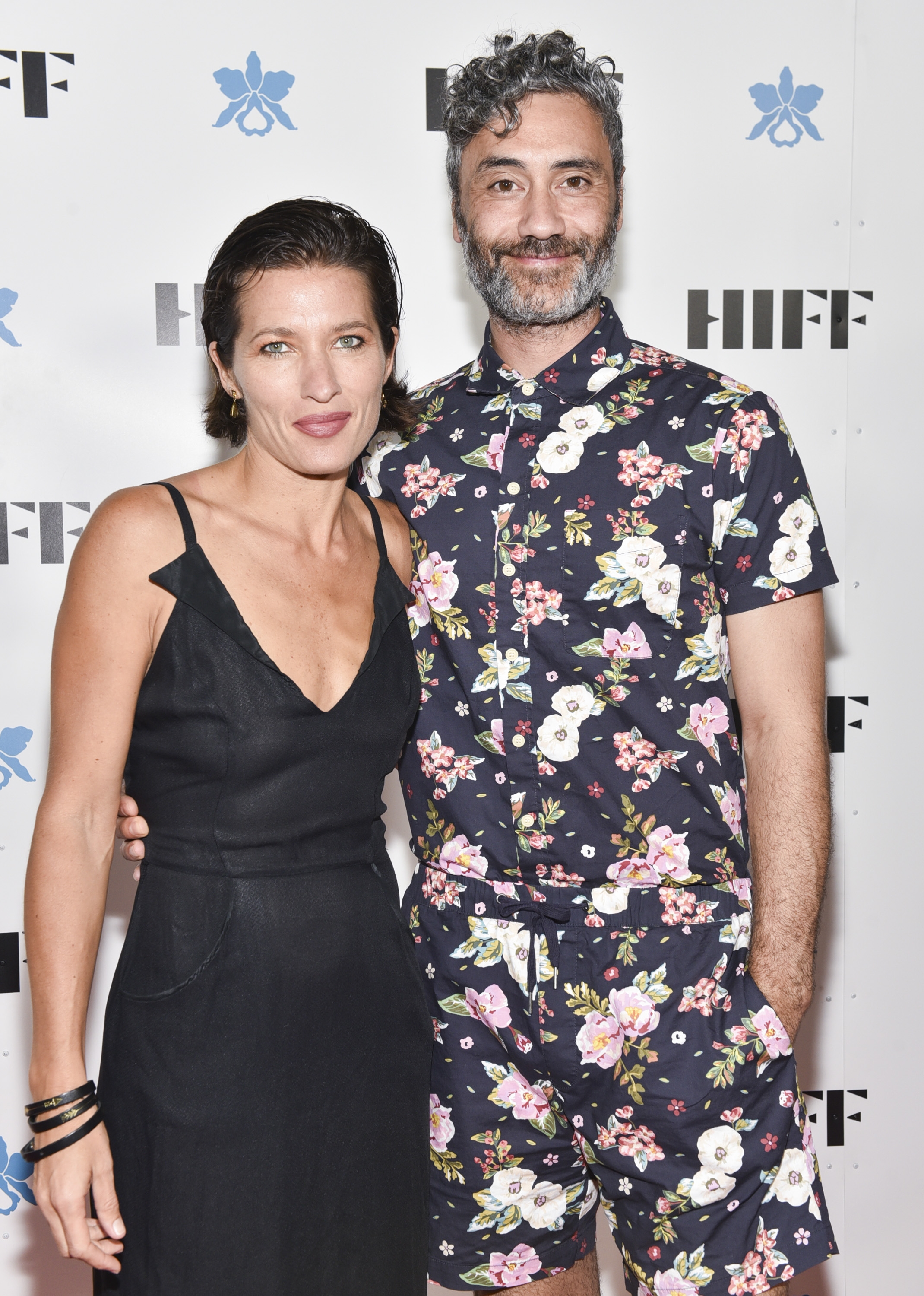 Two people pose together. One wears a simple black dress, the other a floral-patterned shirt and shorts