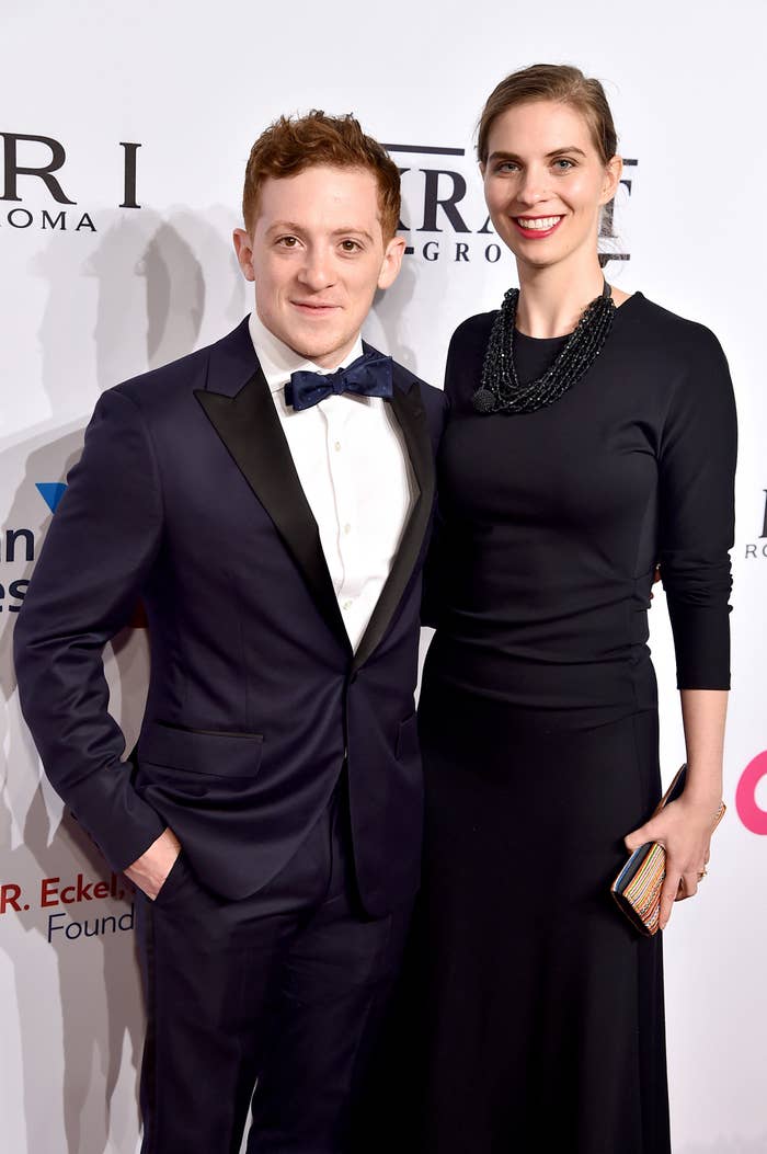 A man in a tuxedo and a woman in an elegant black dress pose together on a red carpet event