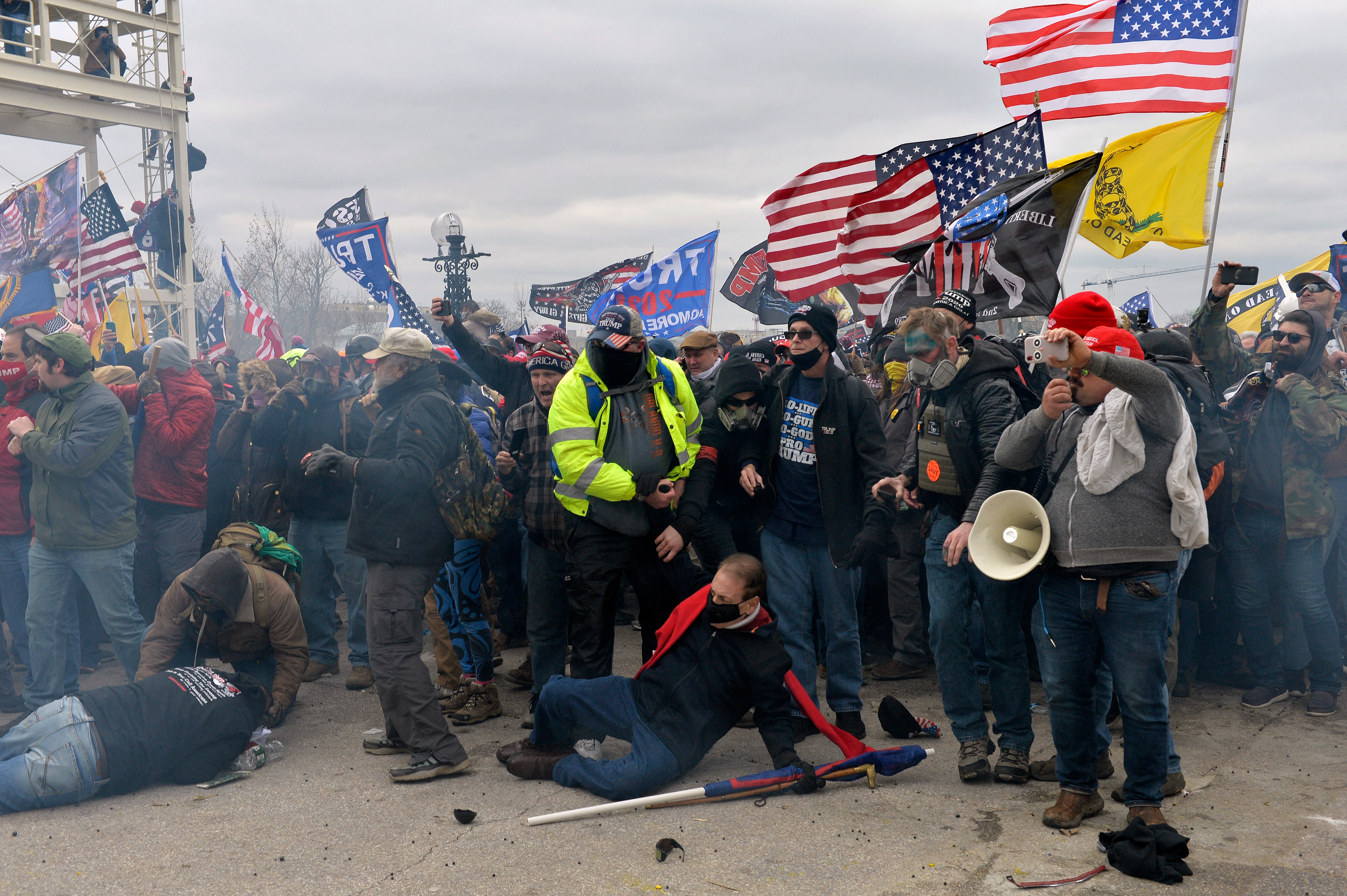 Crowd gathers near flags and banners, some in winter clothes. A person kneels on the ground. The scene appears tense and crowded