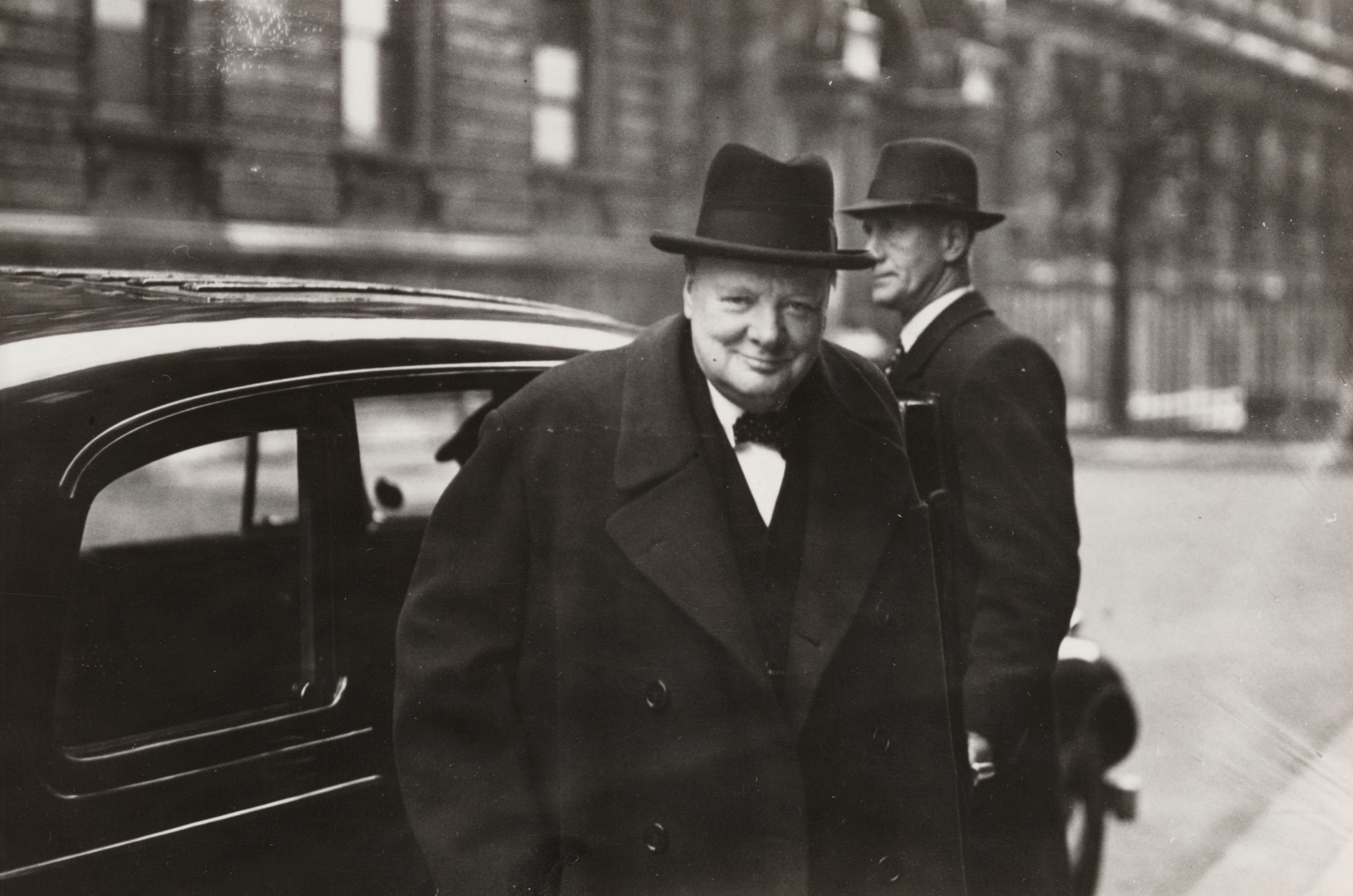 Smiling man in a formal overcoat and bowler hat stands by a vintage car, another man in similar attire behind him on a city street
