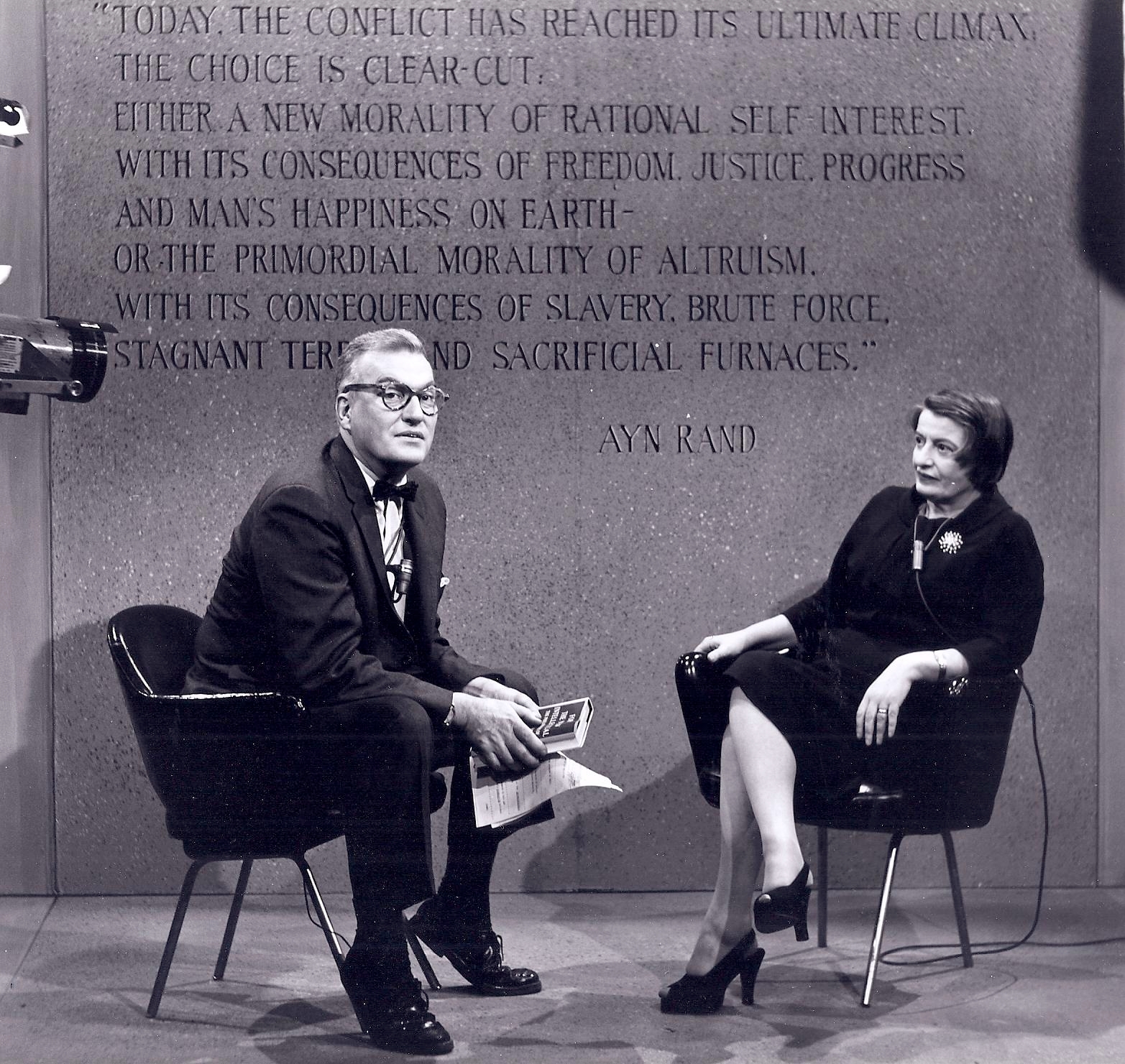 A man and woman seated in discussion on a TV set, with a backdrop featuring a quote by Ayn Rand